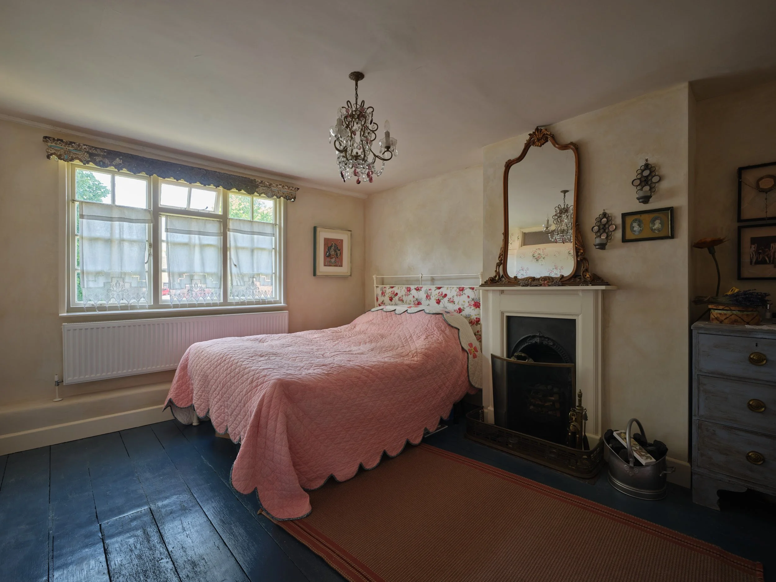 A bedroom with a pink quilted bedspread, floral headboard, and vintage décor including a mirror above a fireplace, chandelier, and framed art on cream-colored walls, wooden floors, and natural light from a window.