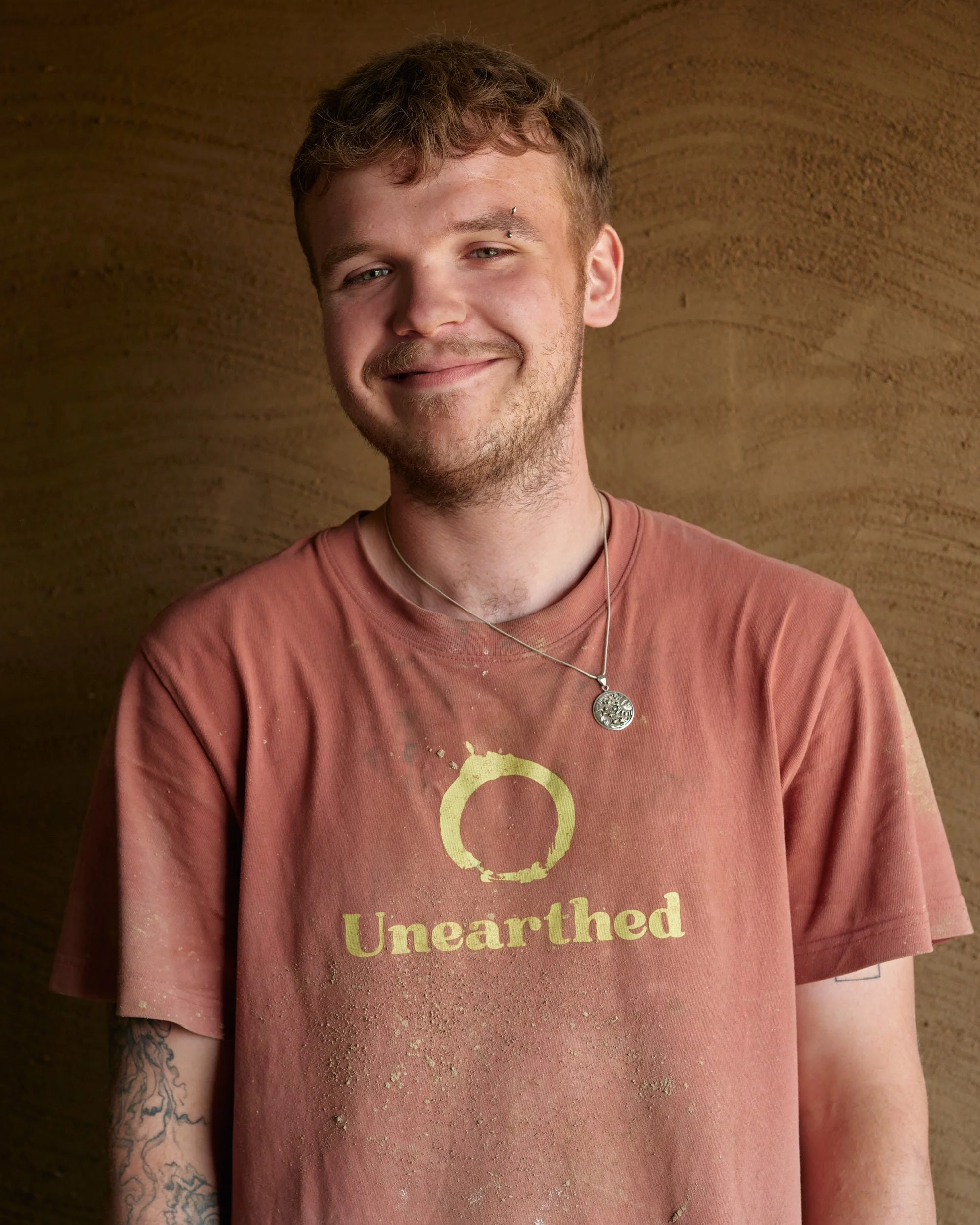 A young man with short curly hair laughing, wearing a brown polo shirt with white paint splatters, sitting against a plain background.
