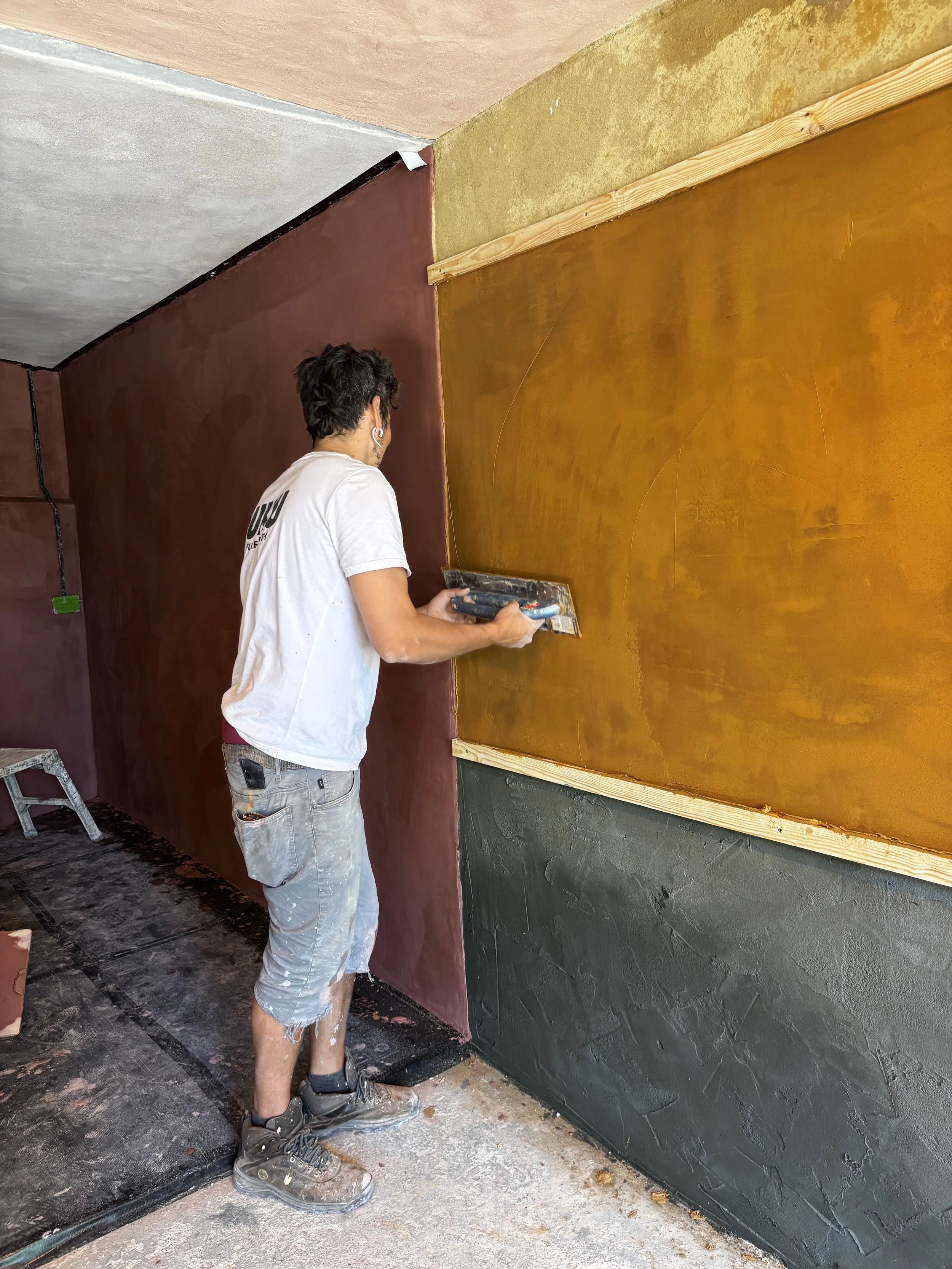 A man wearing a white t-shirt and gray shorts is painting a wall in a room. The wall is divided into sections painted in dark red, mustard yellow, and dark green. The man is using a roller brush and standing on a dusty floor.