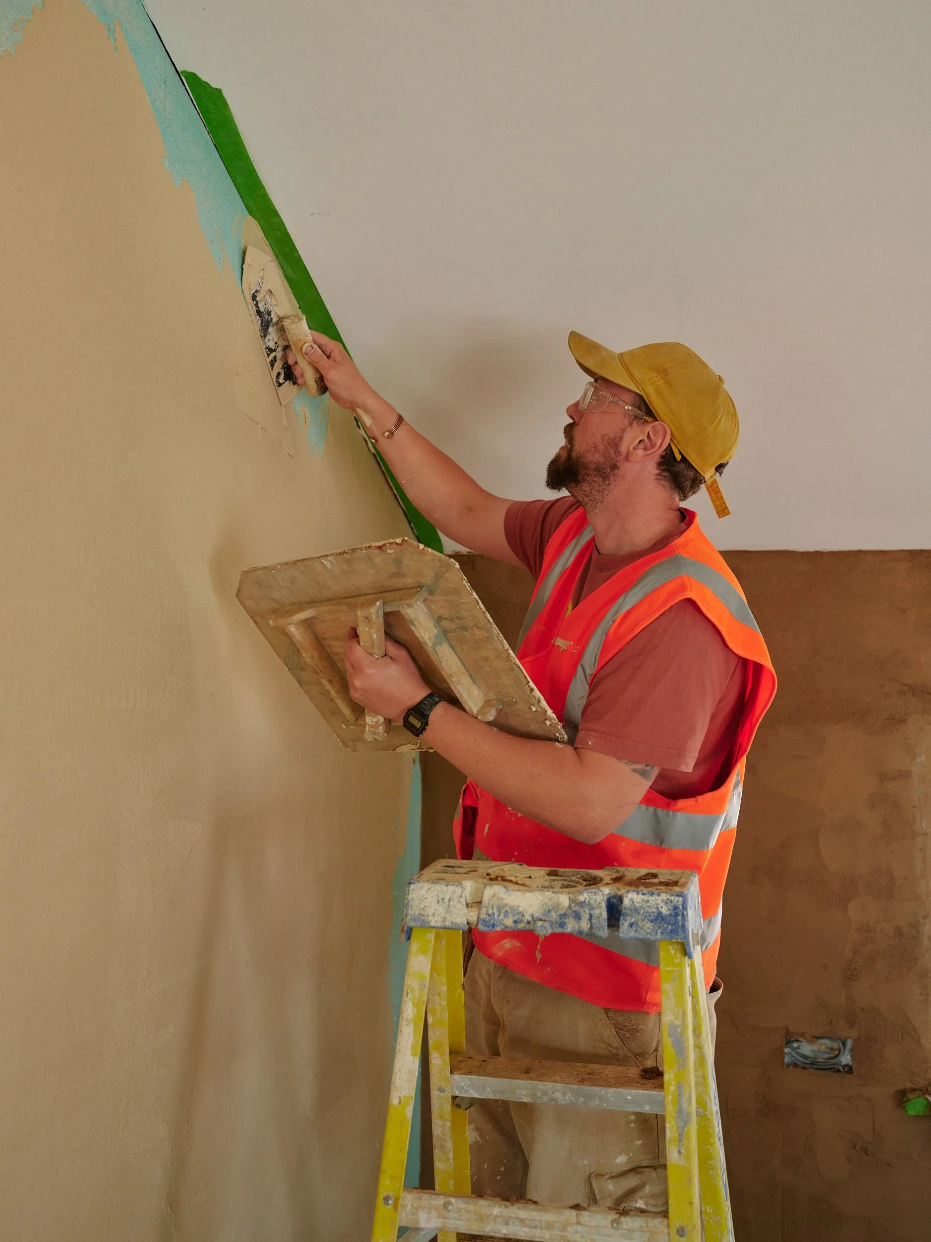 A man wearing a yellow cap, glasses, a red T-shirt, and a bright orange safety vest is standing on a yellow step ladder painting a wall. He is using a paint roller on a section of the wall near the ceiling.