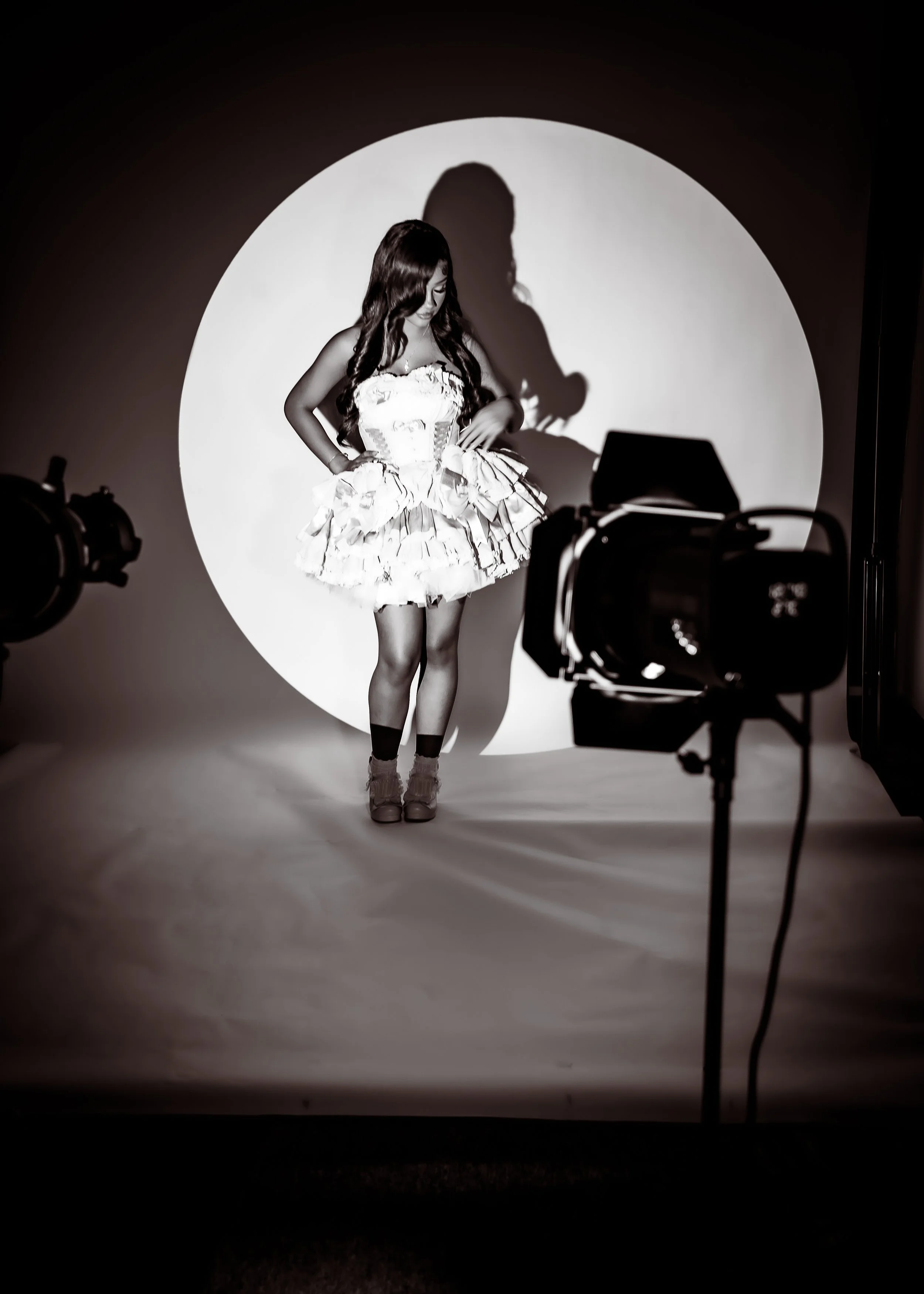 A woman with long hair wearing a ruffled dress and high heels standing in front of a circular spotlight with a camera set up in front of her.