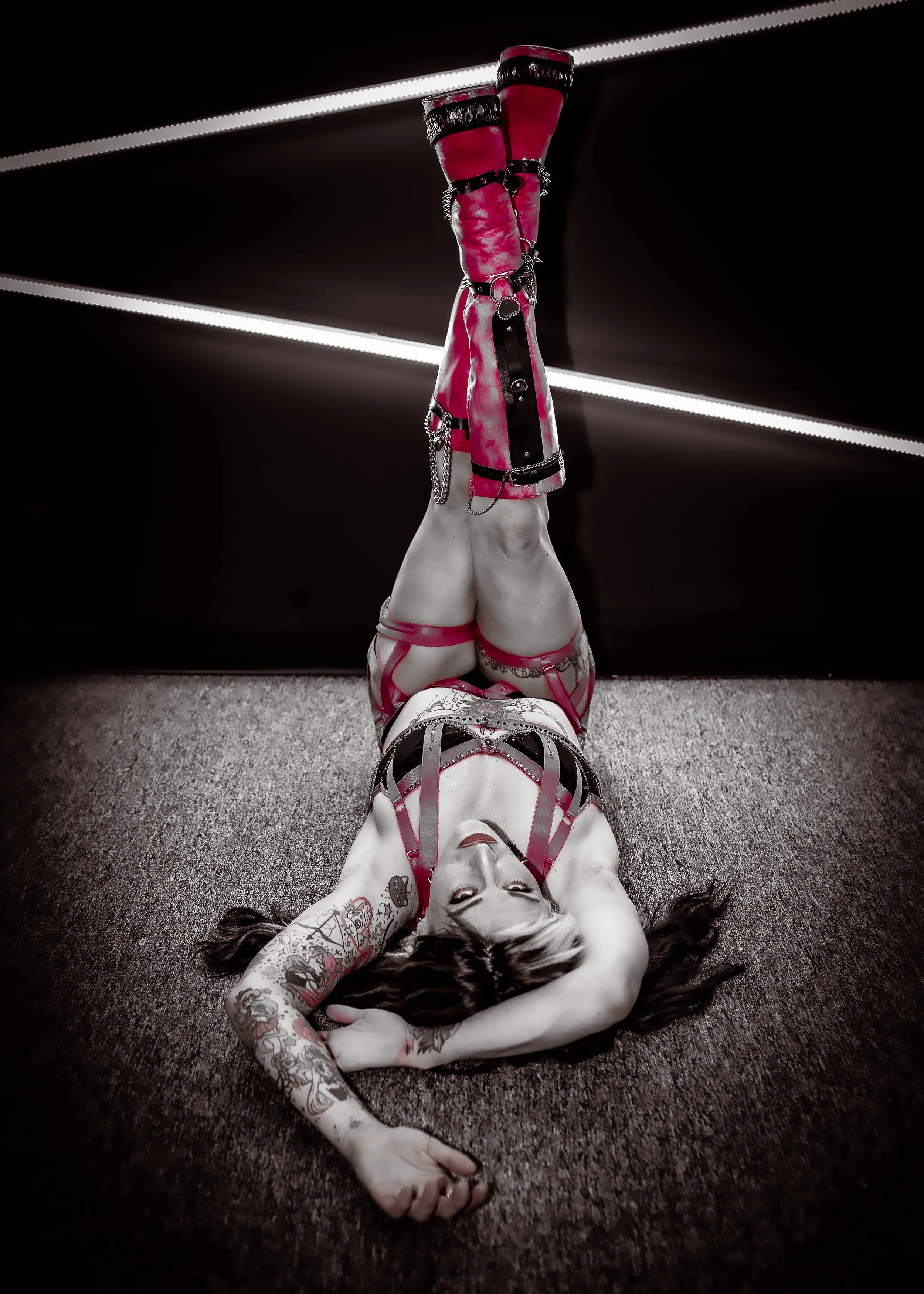 A woman with tattoos lying on the floor with her legs raised and resting against a dark wall with horizontal white lights, wearing pink and black bondage-style outfit and footwear.