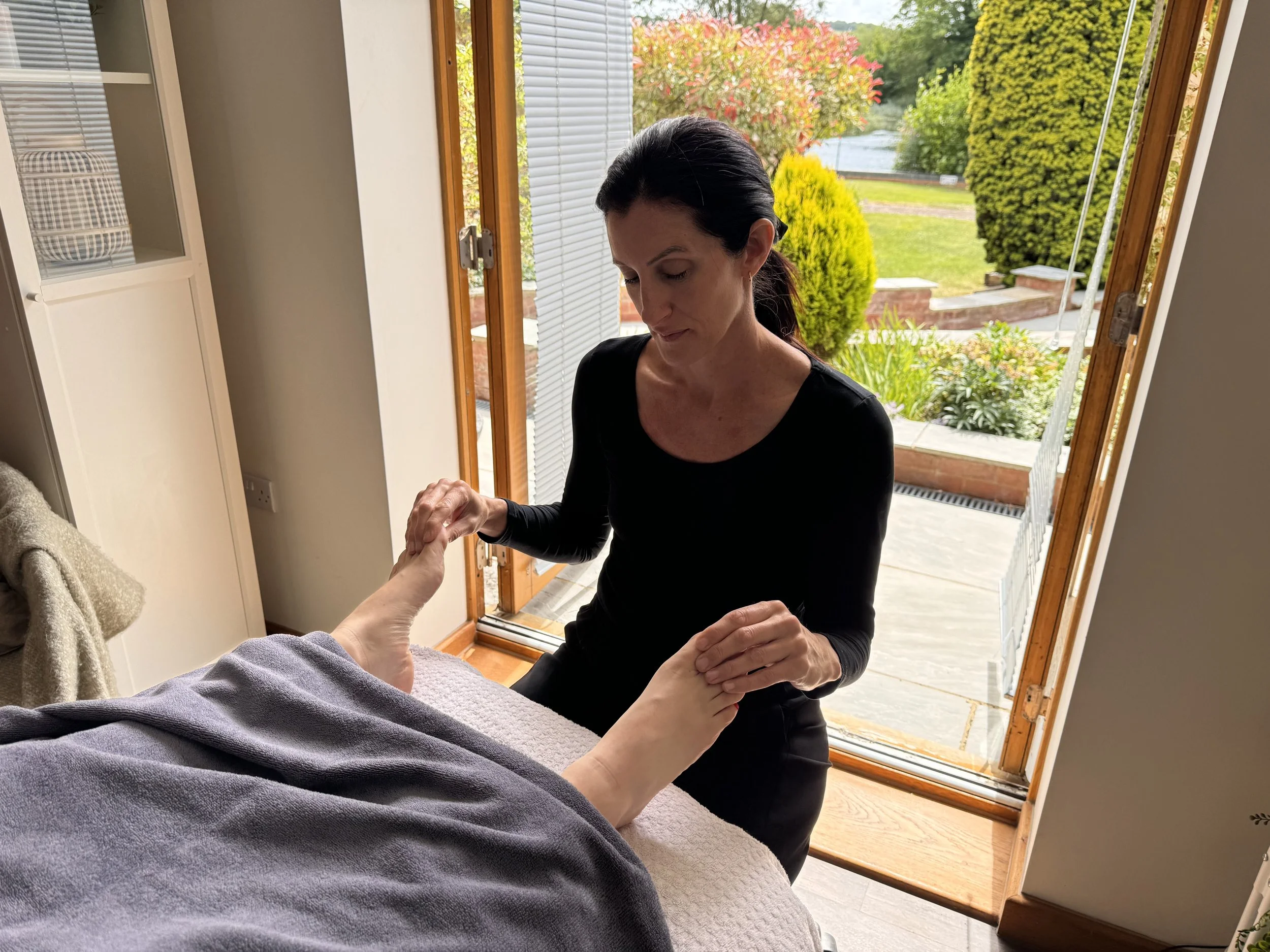 A person in a dark shirt performing a foot massage on someone covered with a blanket in a bright room with view of garden.