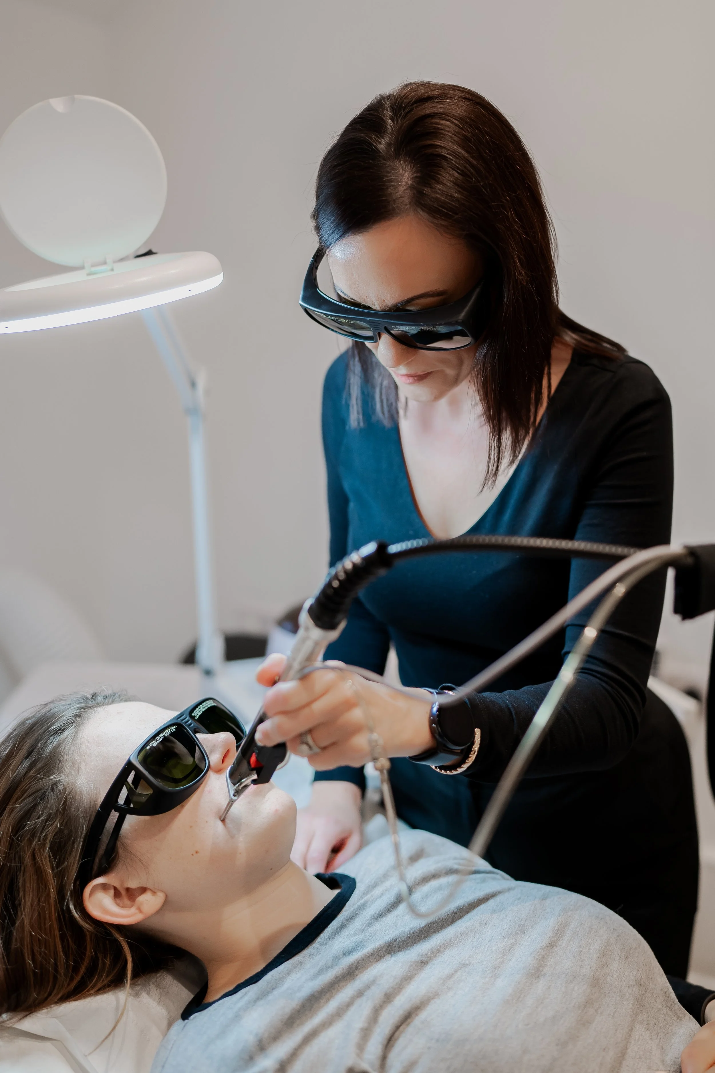 Woman receiving laser treatment, both wearing protective goggles, in a clinic setting.