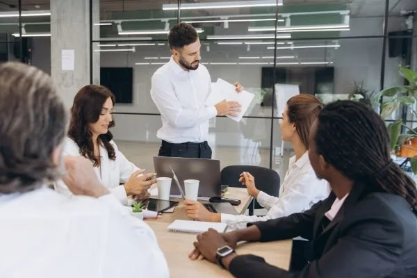 A businessman standing at a conference table, presenting printed documents to a team of colleagues during an office meeting