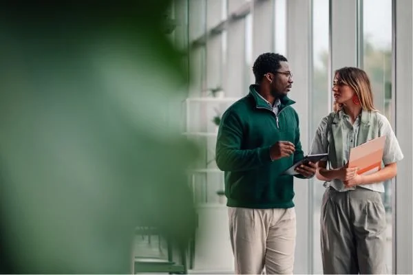 Two colleagues walking alongside a floor-to-ceiling glass window while holding a tablet and folder