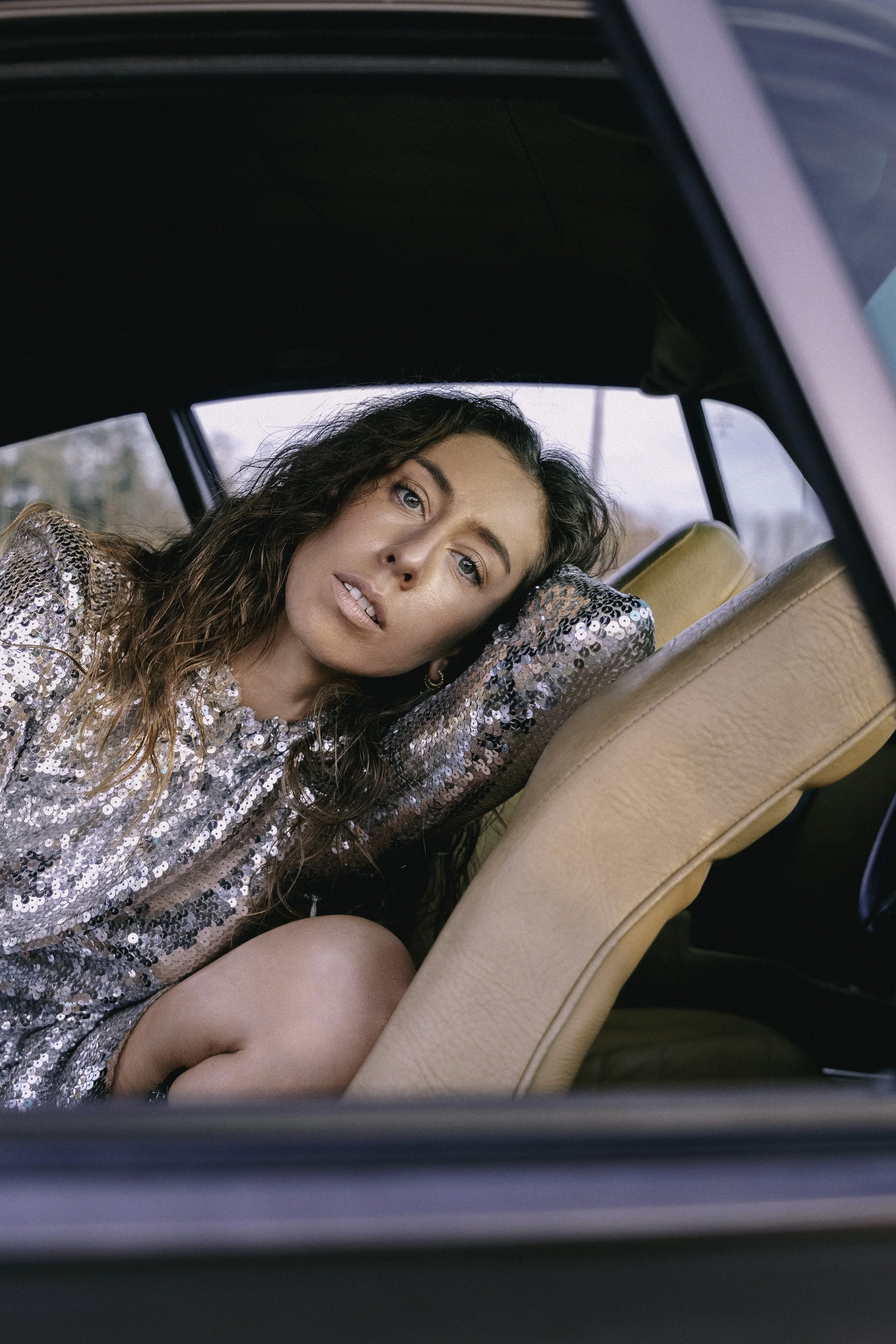 Jeune femme aux cheveux ondulés, portant une robe ou un haut pailleté, assise dans une voiture, posant avec la tête appuyée sur sa main, regardant vers l'objectif.