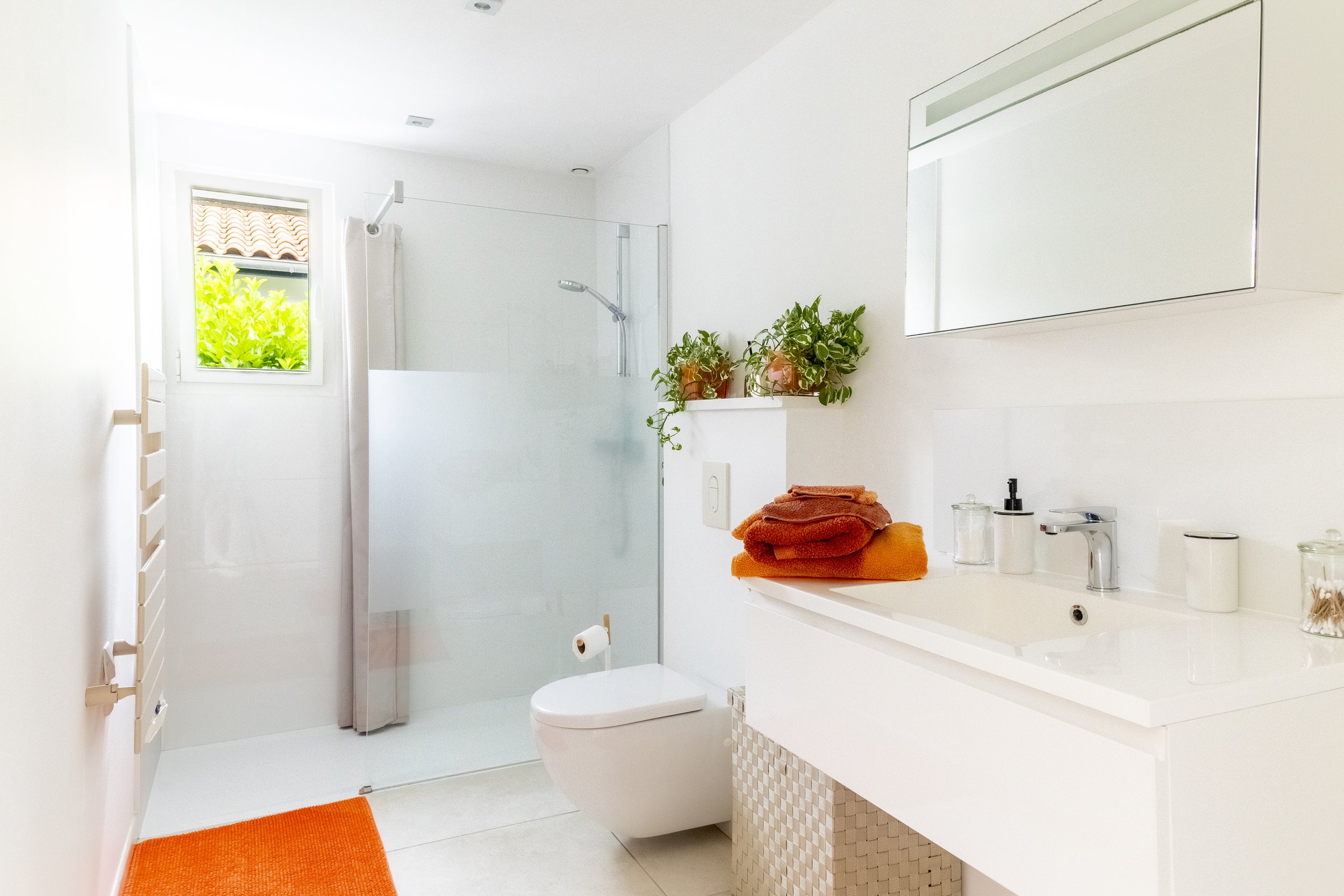 Une salle de bain moderne et lumineuse avec un mur blanc, un lavabo avec un miroir, des serviettes orange pliées sur le plan de toilette, des plantes dans des pots, une fenêtre avec vue sur un extérieur verdoyant, et un espace douche avec une porte en verre.