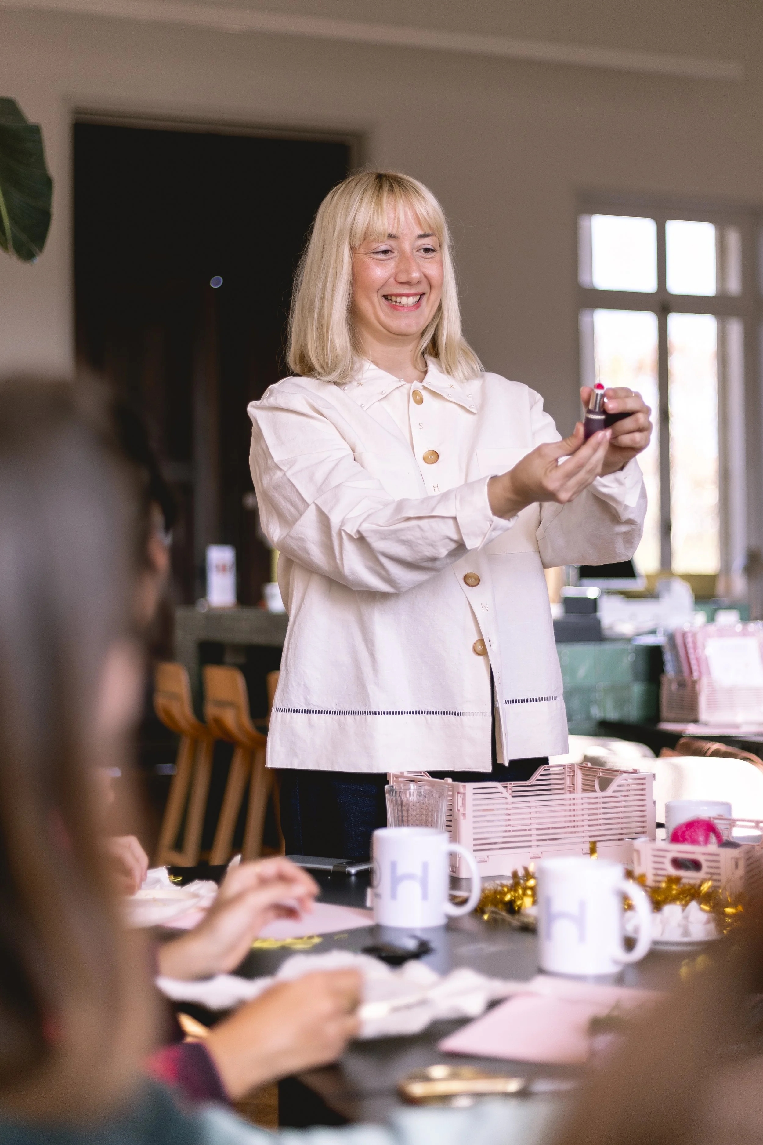 Une femme souriante, blonde, habillée d'une blouse claire, regardant un rouge à lèvres dans une pièce lumineuse avec une fenêtre en arrière-plan, lors d'une réunion ou d'un événement festif.