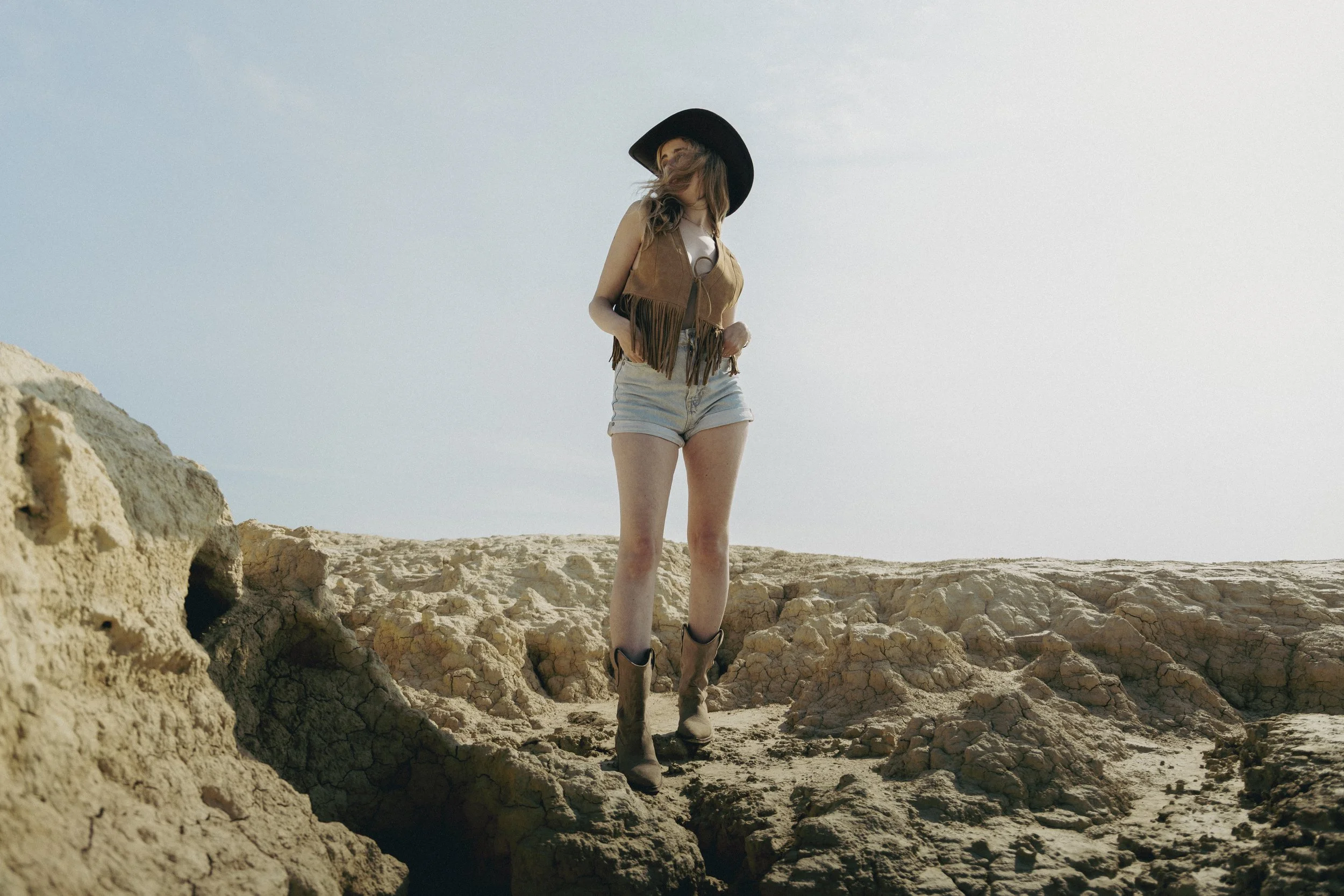 Jeune femme portant un chapeau noir, une veste en cuir avec franges, un t-shirt blanc, des shorts en denim, et des bottes de cow-boy, se tenant sur un terrain rocheux dans un paysage désertique.