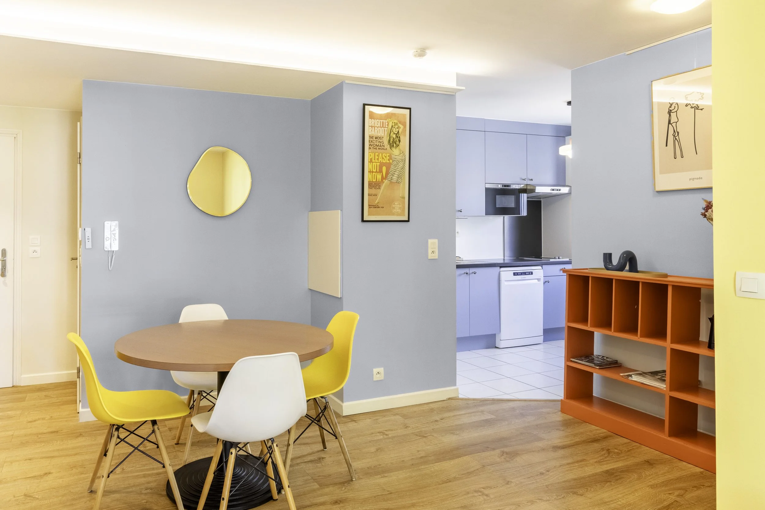 Salle à manger avec une table ronde en bois entourée de chaises de couleurs différentes, murs bleu pâle, cuisine en arrière-plan avec armoires bleues, radiateur orange, art mural, miroir, et une petite étagère orange.