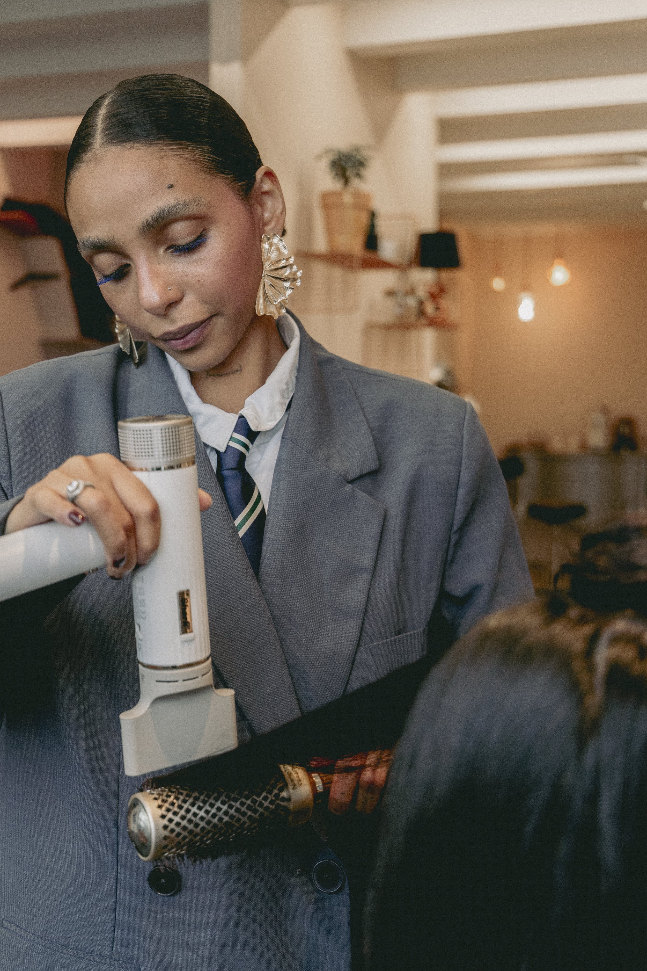 Une femme portant un tailleur gris foncé et une cravate, tenant un objet cylindrique avec un pinceau à maquillage ou un outil cosmétique, dans un intérieur chaleureux avec des lampes suspendues et des étagères en bois.