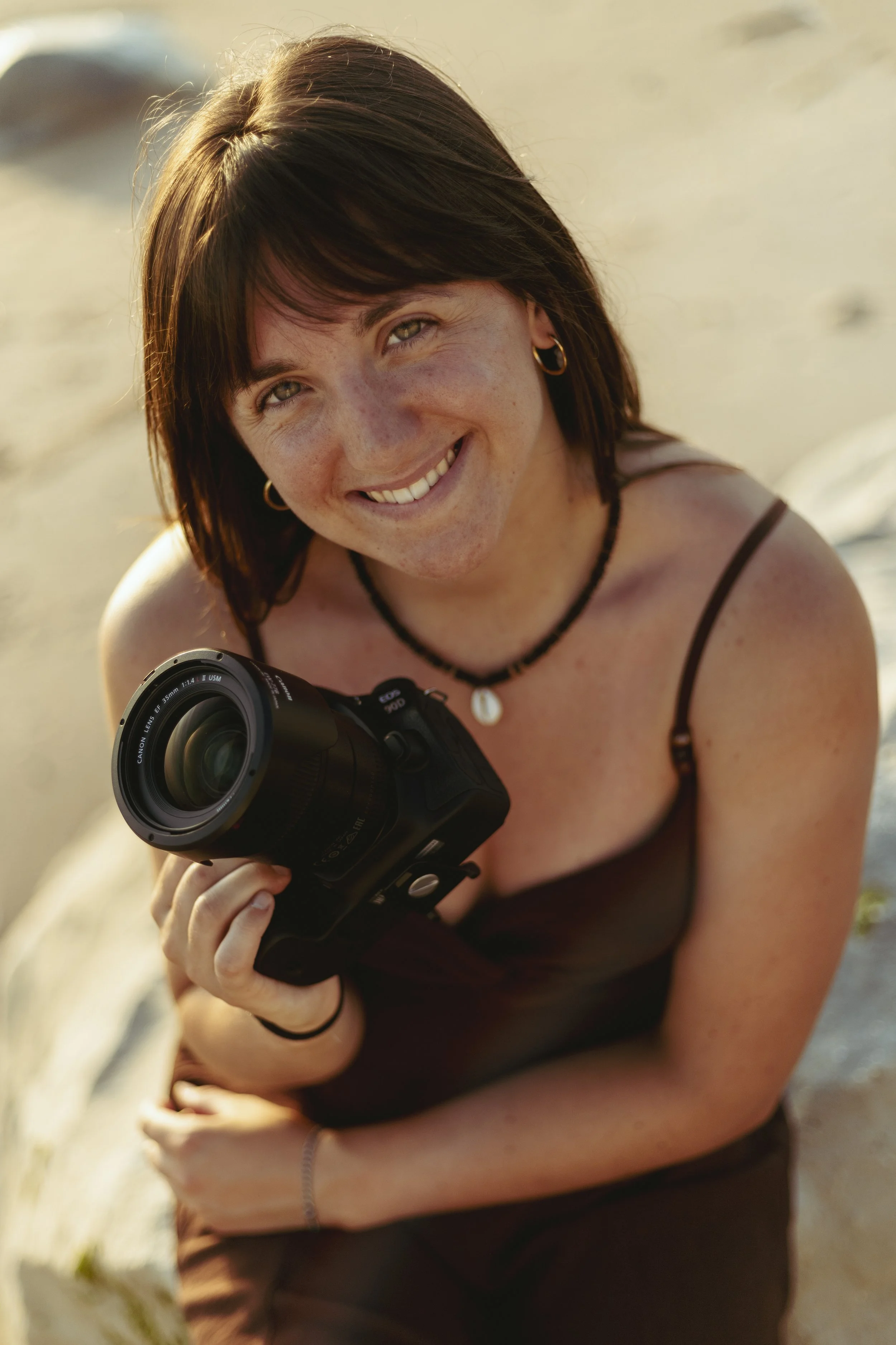 Une femme souriante tenant un appareil photo numérique, avec un fond de plage ensoleillé.