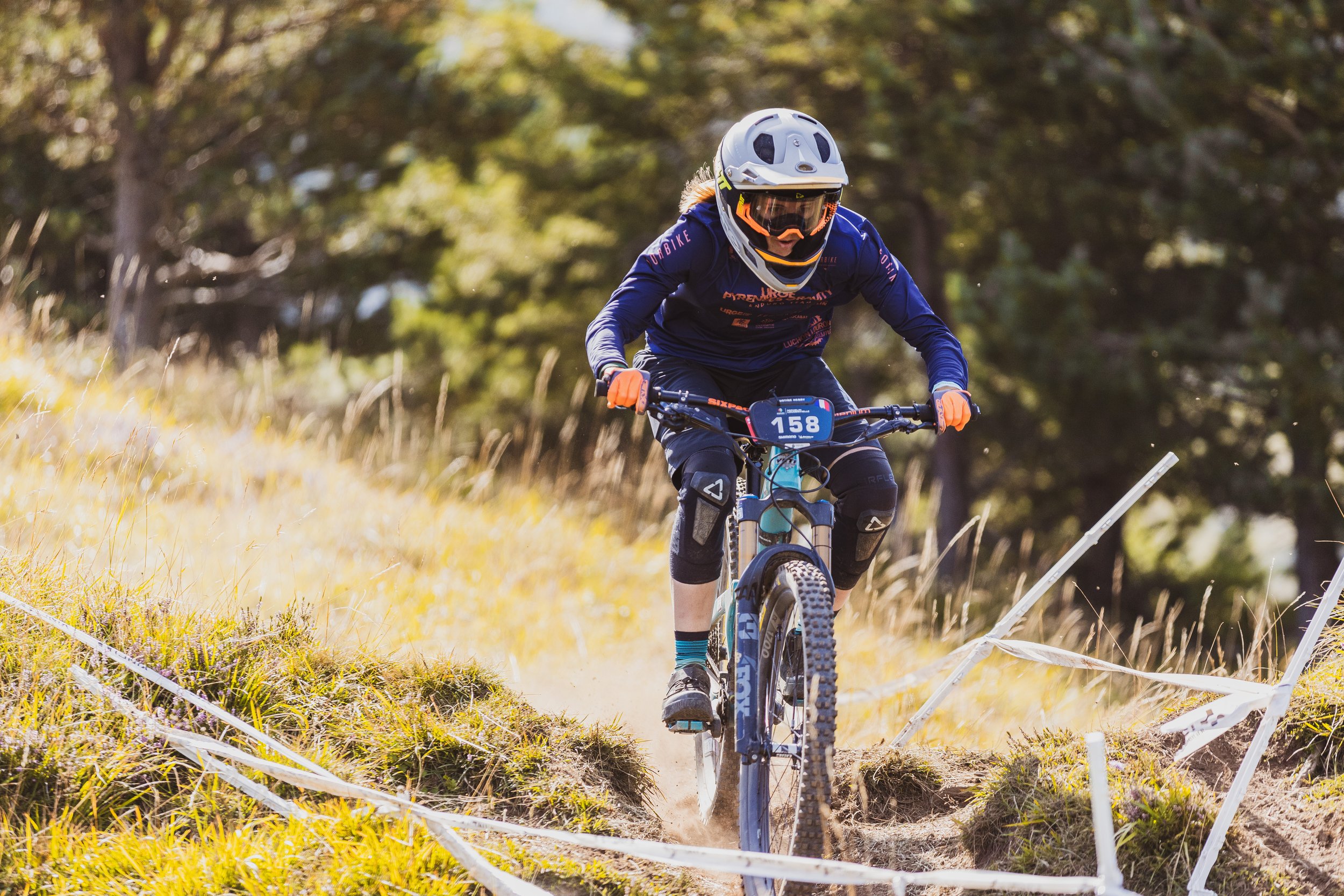 Coureur de VTT portant un casque, des lunettes de protection et un maillot bleu, pédalant sur une piste en nature entourée d'herbe et d'arbres, avec des banderoles blanches délimitant la course.
