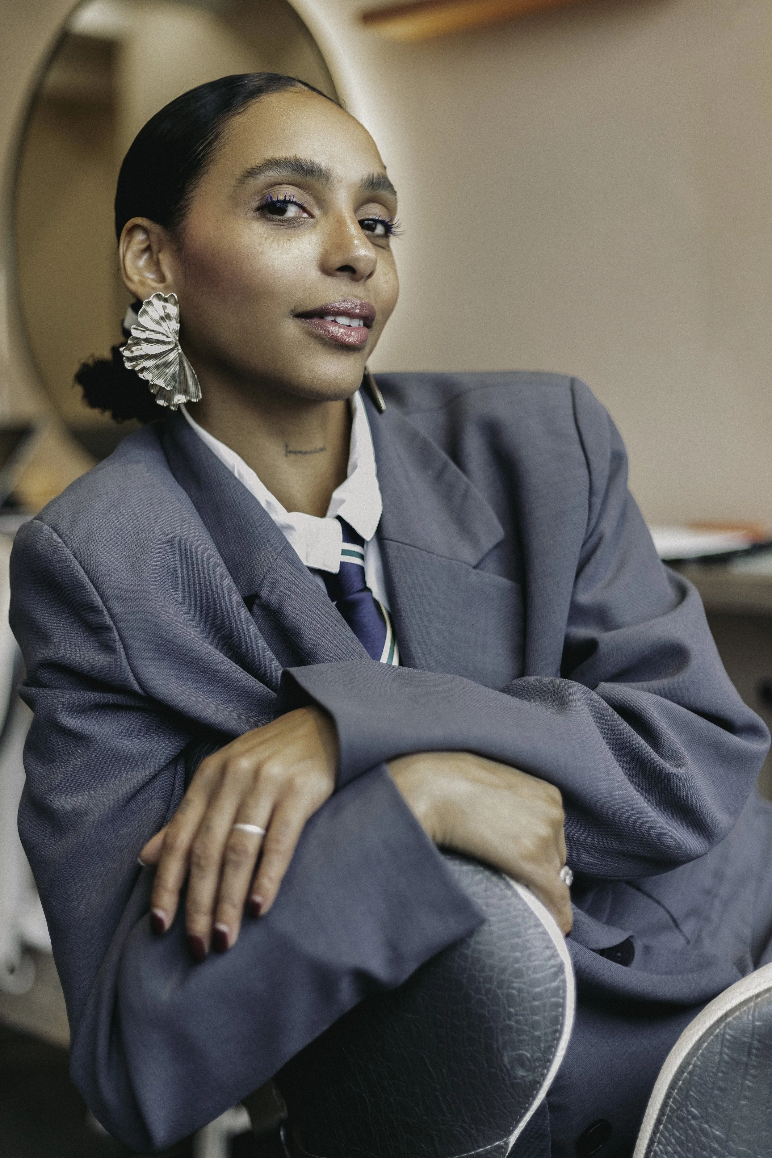 Une femme portant un costume gris foncé avec une chemise blanche et une cravate, assise dans une salle, avec ses bras croisés sur ses jambes, portant de grandes boucles d'oreilles en forme de feuille et un maquillage léger, souriante légèrement. En a