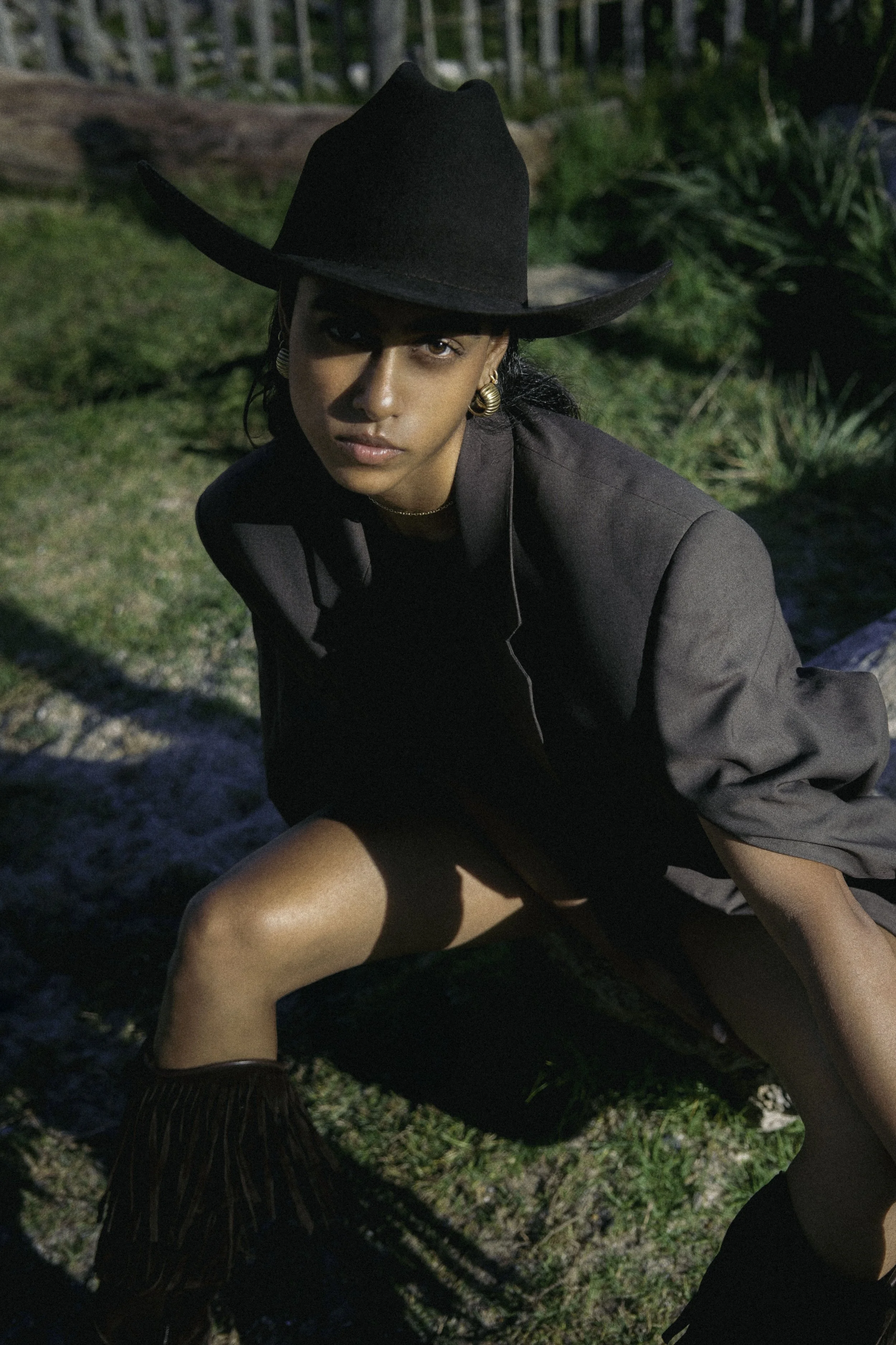 Femme portant un chapeau noir, une veste grise, et des bottines à franges, posant en extérieur.