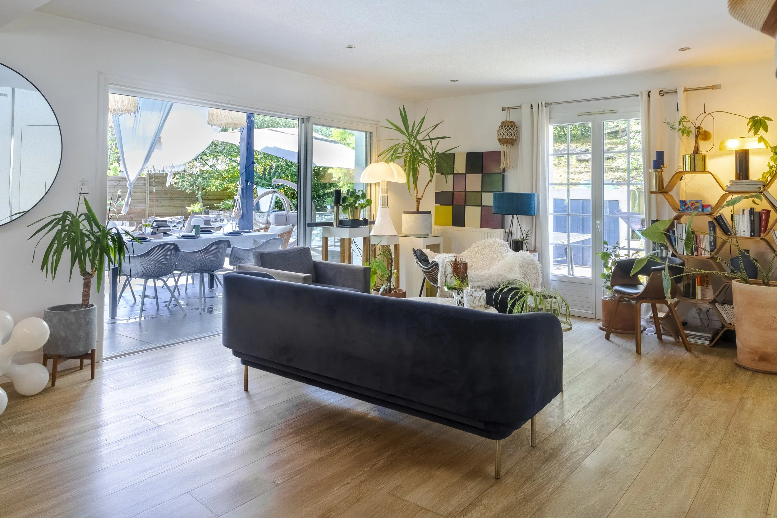 Salon avec canapé noir, plusieurs plantes vertes, grande fenêtre avec vue sur jardin, table à manger extérieure, décorations modernes.