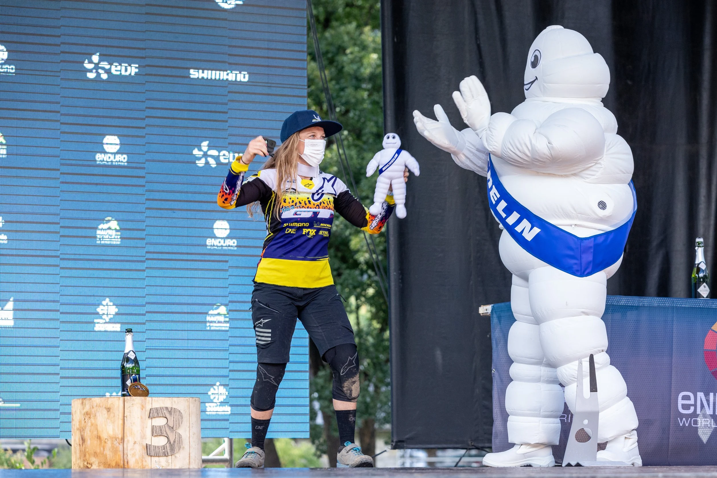 Une femme en tenue de sport et portant un masque facial, en train de se prendre en photo avec une mascotte Michelin lors d'un événement en plein air.