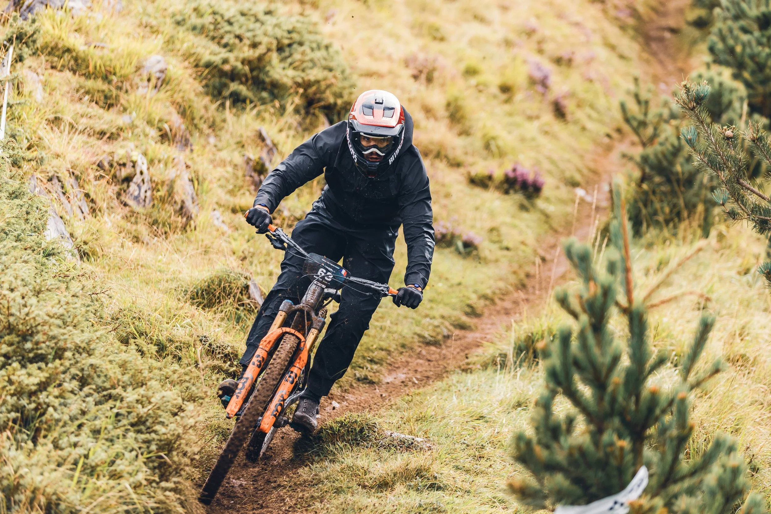 Un homme en équipement de protection fait du VTT sur un sentier en pleine nature, entouré de végétation dense et de petites montagnes.