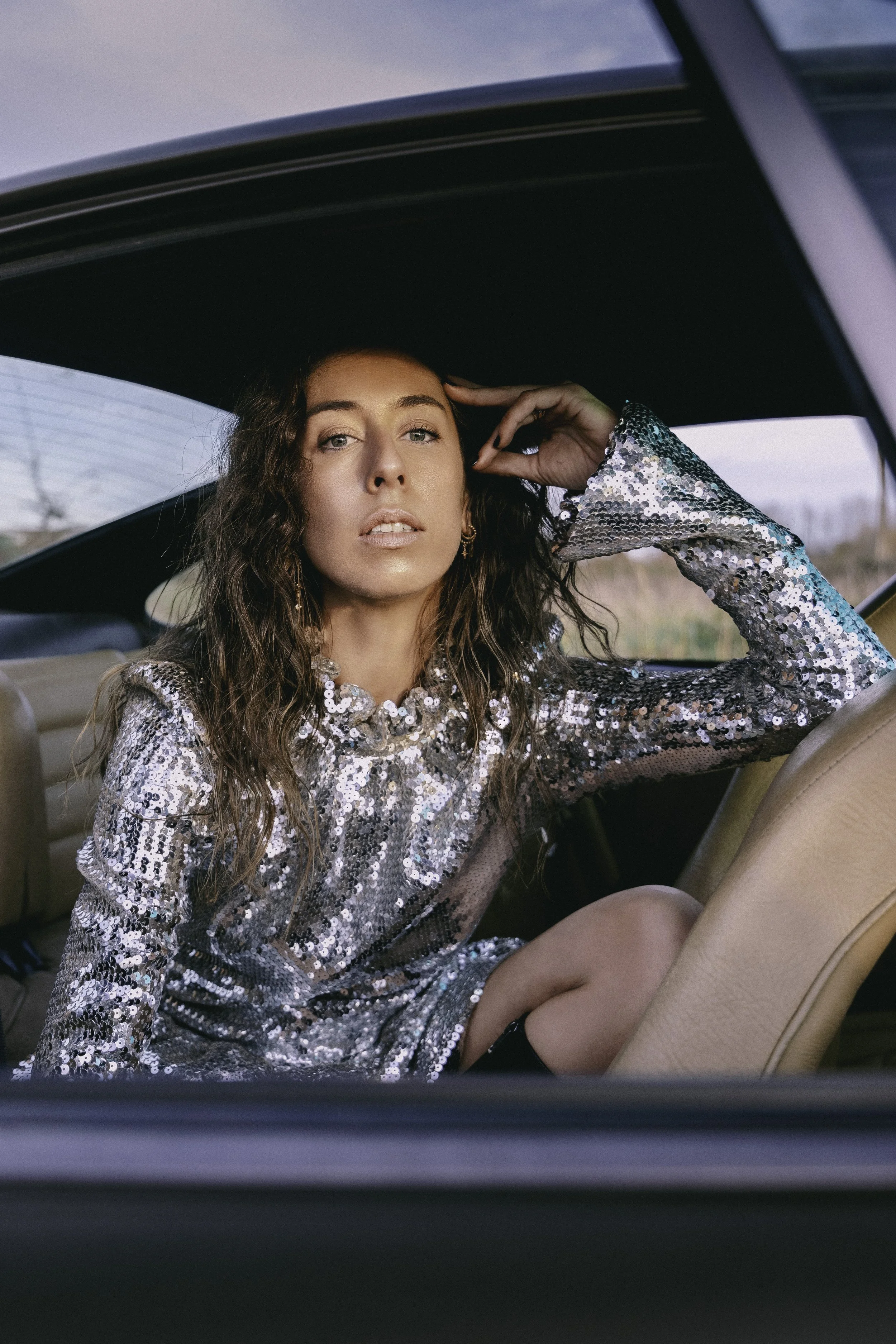 Une femme avec des cheveux ondulés portant une robe à paillettes argentées, assise dans une voiture, regardant vers l'extérieur.