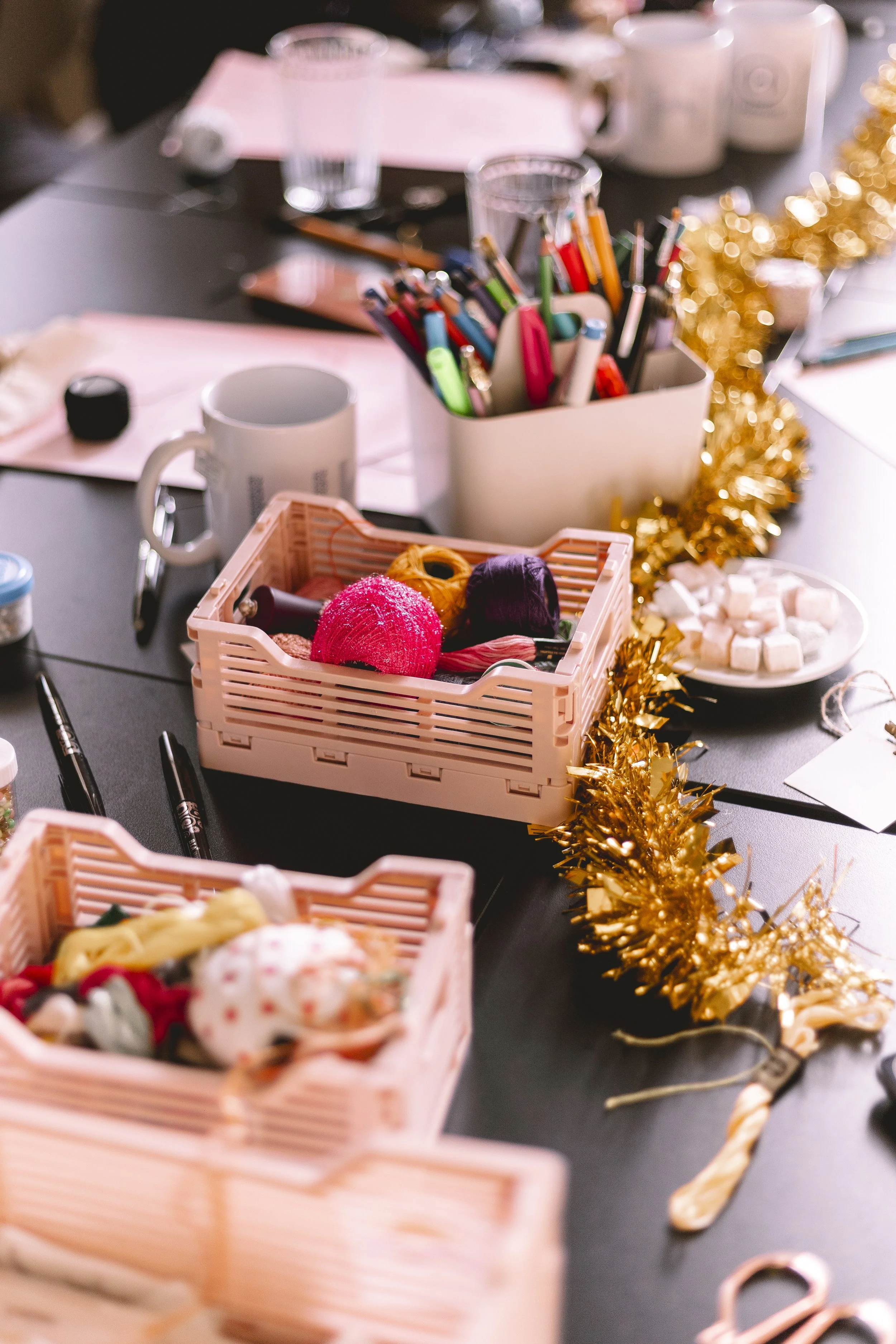 Un bureau décoré avec une guirlande dorée, des stylos, des rubans, des bonbons, des pelotes de fil de différentes couleurs, une tasse blanche, et d'autres objets de bureau et décorations festives.