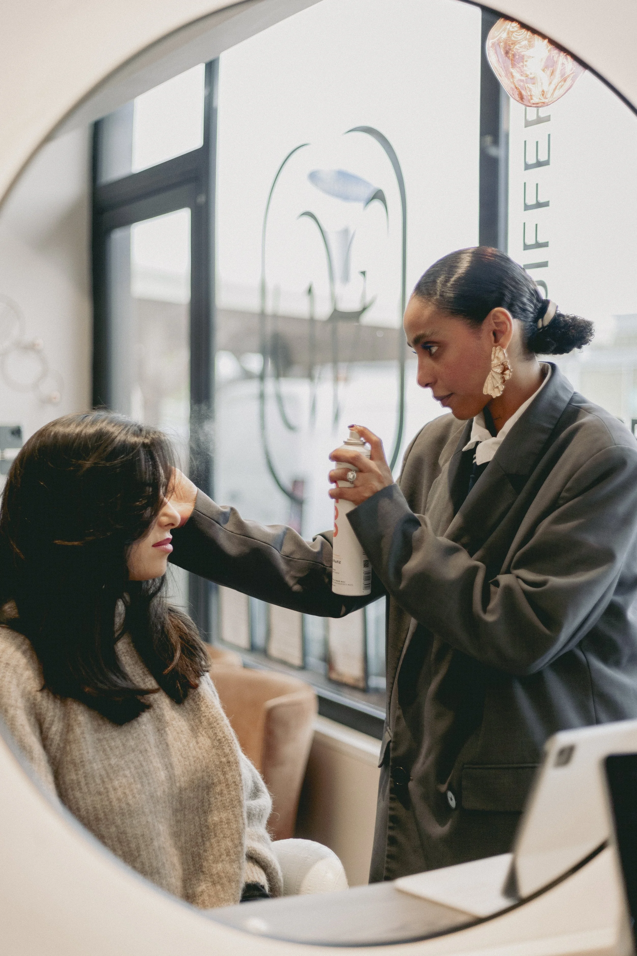 Vue à travers une ouverture ronde montrant une femme faisant un maquillage ou un soin à une autre femme dans un salon ou un espace intérieur, avec des grandes fenêtres en arrière-plan.