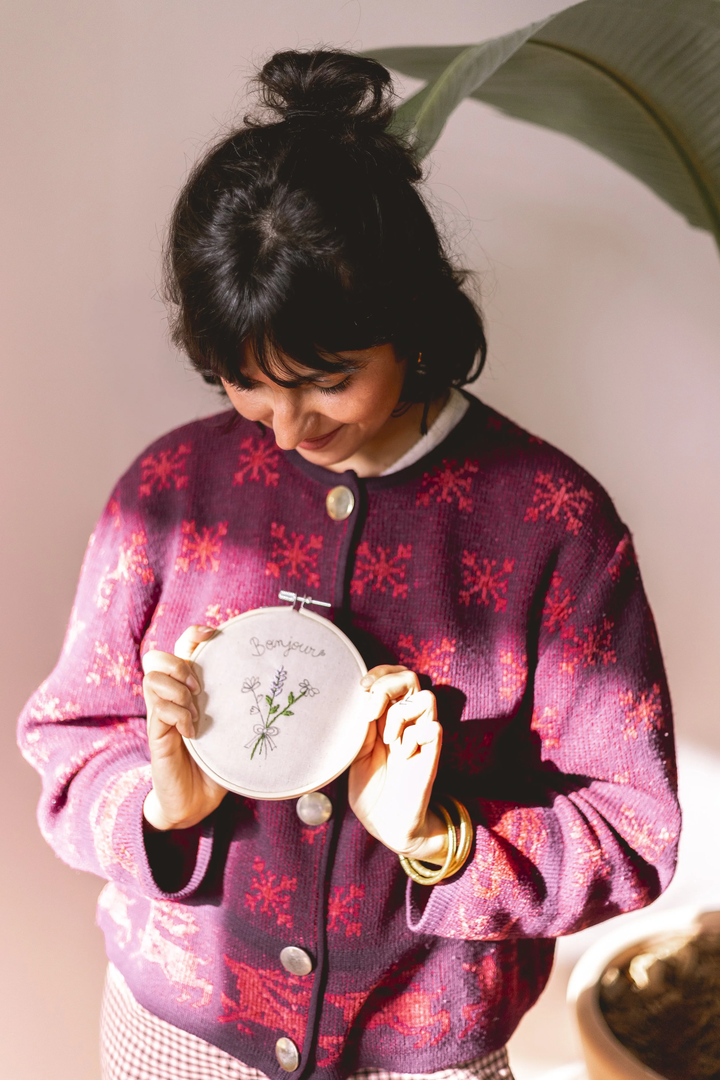 Une femme souriante regarde un cercle de broderie avec le mot 'Bonjour' écrit dessus, entourée de petites fleurs. Elle porte un pull rouge à motifs de flocons de neige et des bracelets en or.