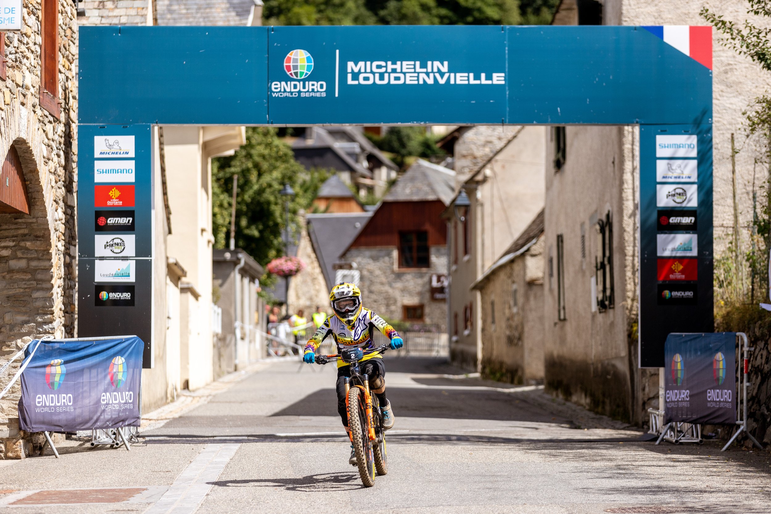 Un cycliste en tenue de course, portant un casque, descend une rue pavée lors d'une compétition de mountain bike Enduro à Loudenvielle, France, sous une arche avec des logos de sponsors.