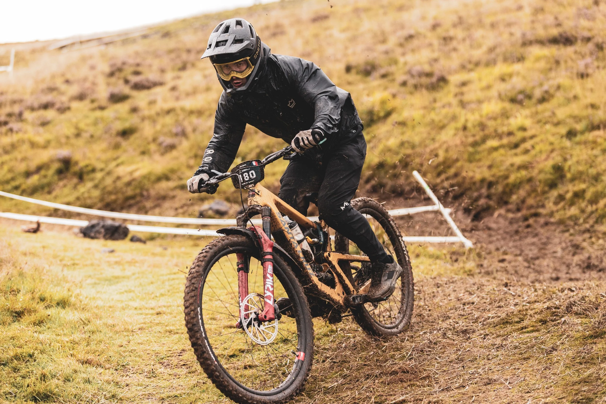 Un cycliste portant un casque et une veste noire, chevauchant un VTT dans un terrain boueux en plein air avec un arrière-plan vallonné.