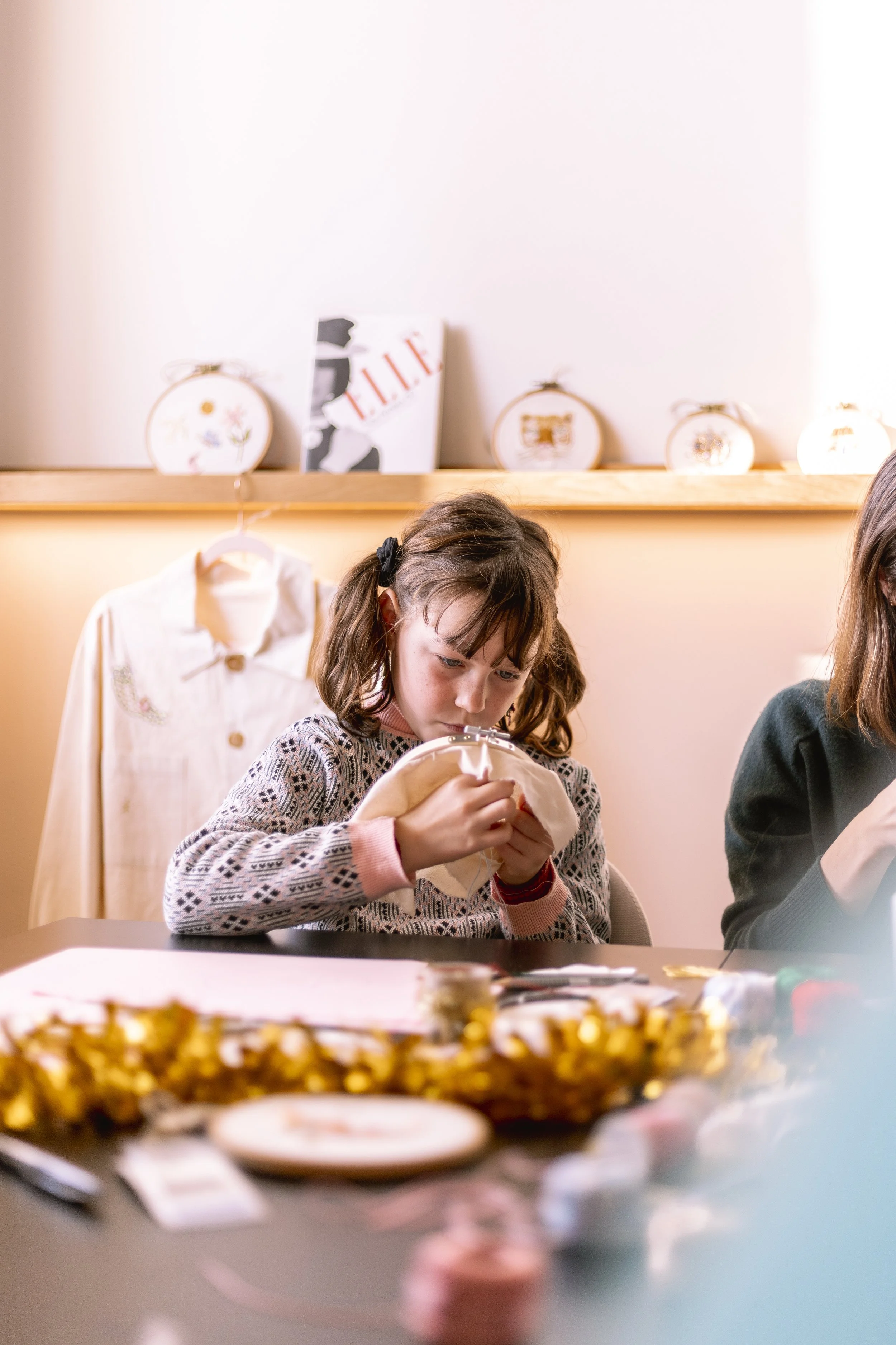 Une jeune fille assise à une table, concentrée sur ses activités. En arrière-plan, décoration de fêtes avec des ballons et un gâteau, ambiance festive.