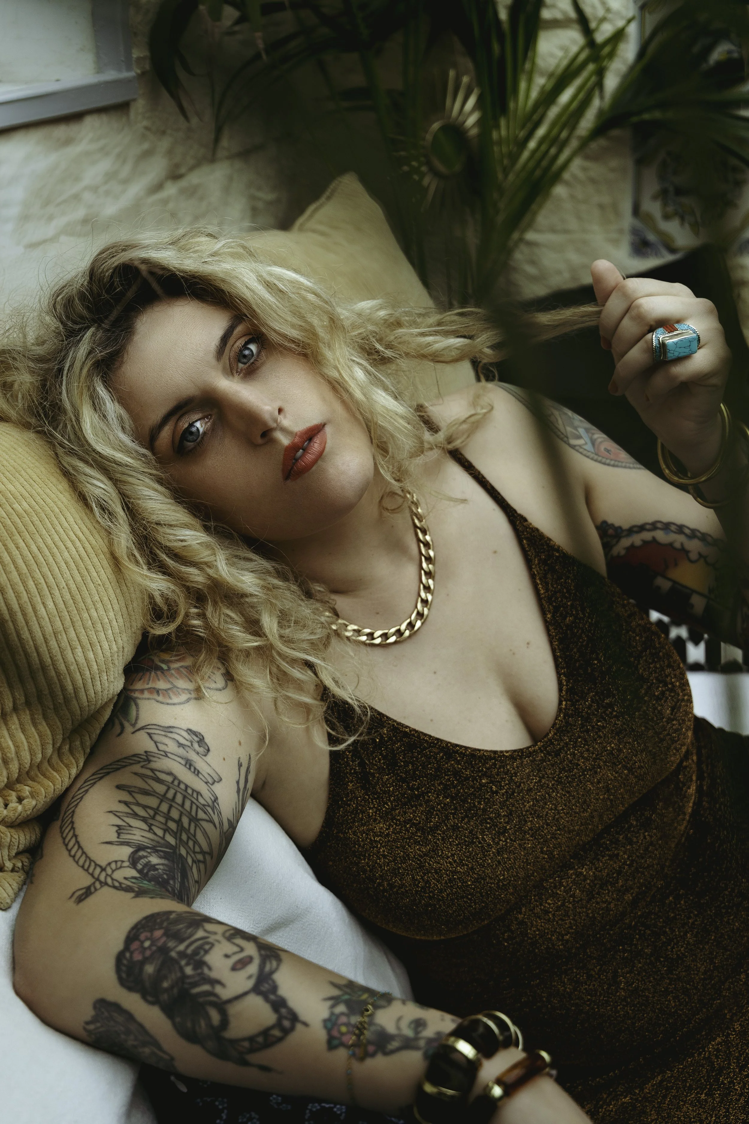 Une femme aux cheveux blonds bouclés repose sur un canapé, vêtue d'une robe moulante à motifs dorés, portant des bijoux en or, un bague turquoise, et affichant plusieurs tatouages, dont une femme avec des fleurs sur le bras. Elle tient une mèche de ses cheveux.