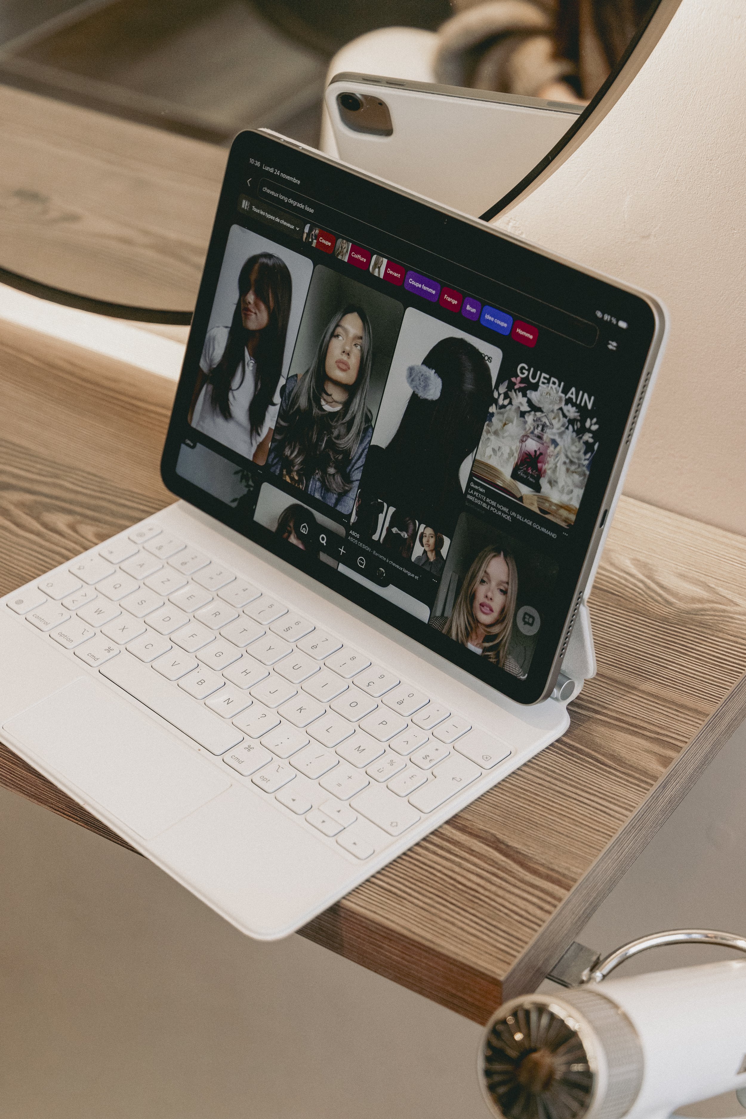 Un ordinateur portable blanc avec écran affichant une page web de mode avec des images de femmes, posé sur une table en bois, avec un miroir rond en arrière-plan et un ventilateur à l'avant.