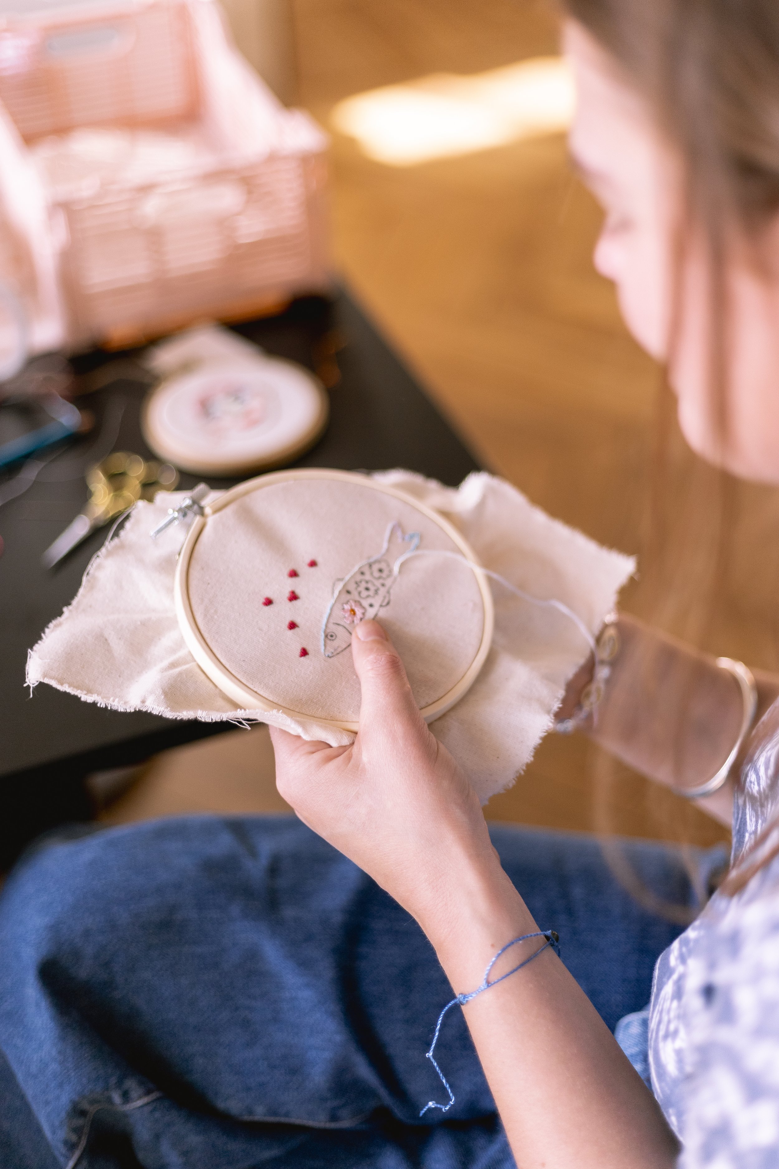 Une personne tenant une boucle de broderie avec une broderie en cours représentant un animal et des petits cœurs rouges, assise avec des jeans bleus, devant une table avec des outils de couture.