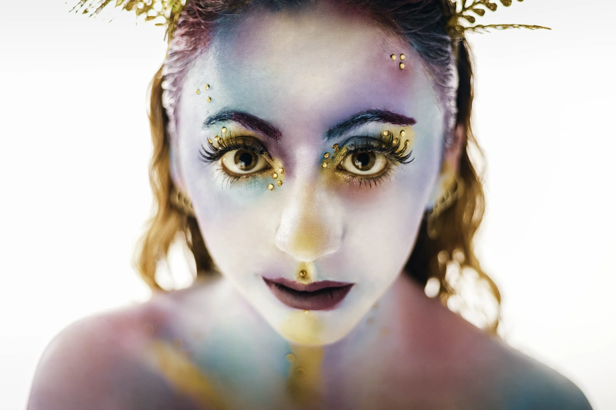 Une personne avec un maquillage coloré et des décorations dorées sur le visage, avec une expression neutre, regardant droit devant, dans un éclairage lumineux.
