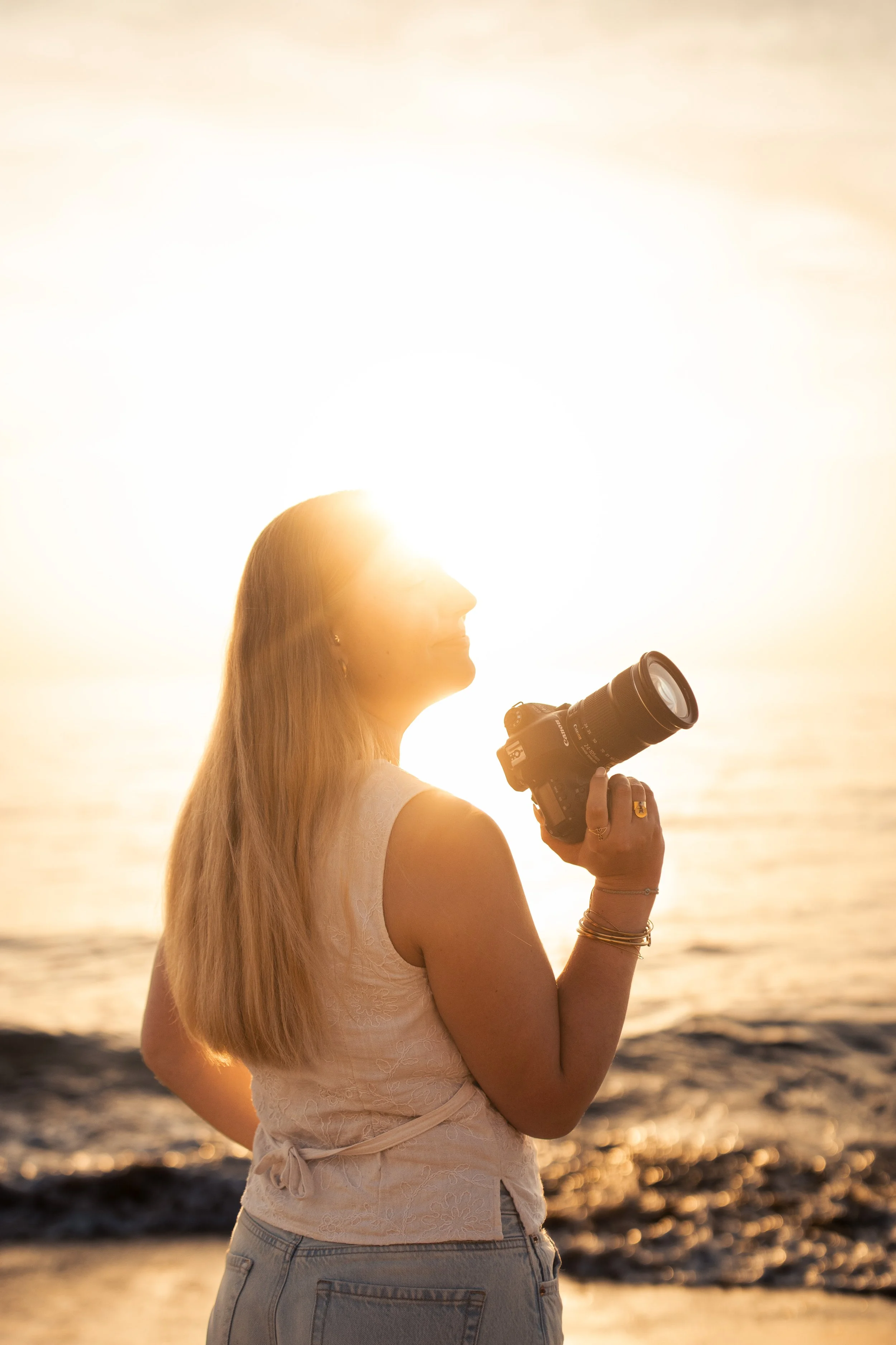 Femme avec des cheveux longs, portant une chemise blanche, tenant un appareil photo lors d'un coucher de soleil à la plage.