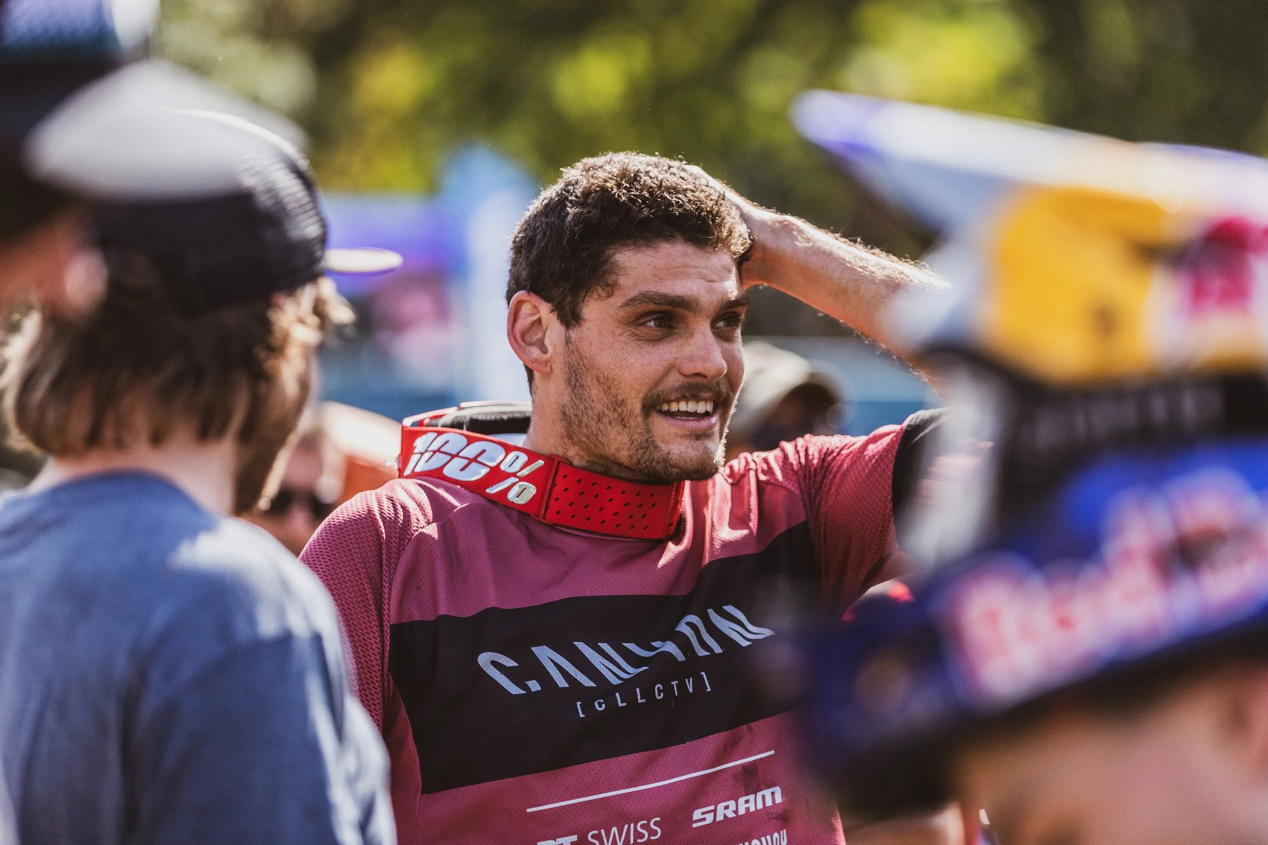 Un jeune homme en maillot de sport rouge et noir célébrant ou discutant avec d'autres personnes lors d'une activité sportive en plein air, avec un fond flou d'arbres et de personnes.