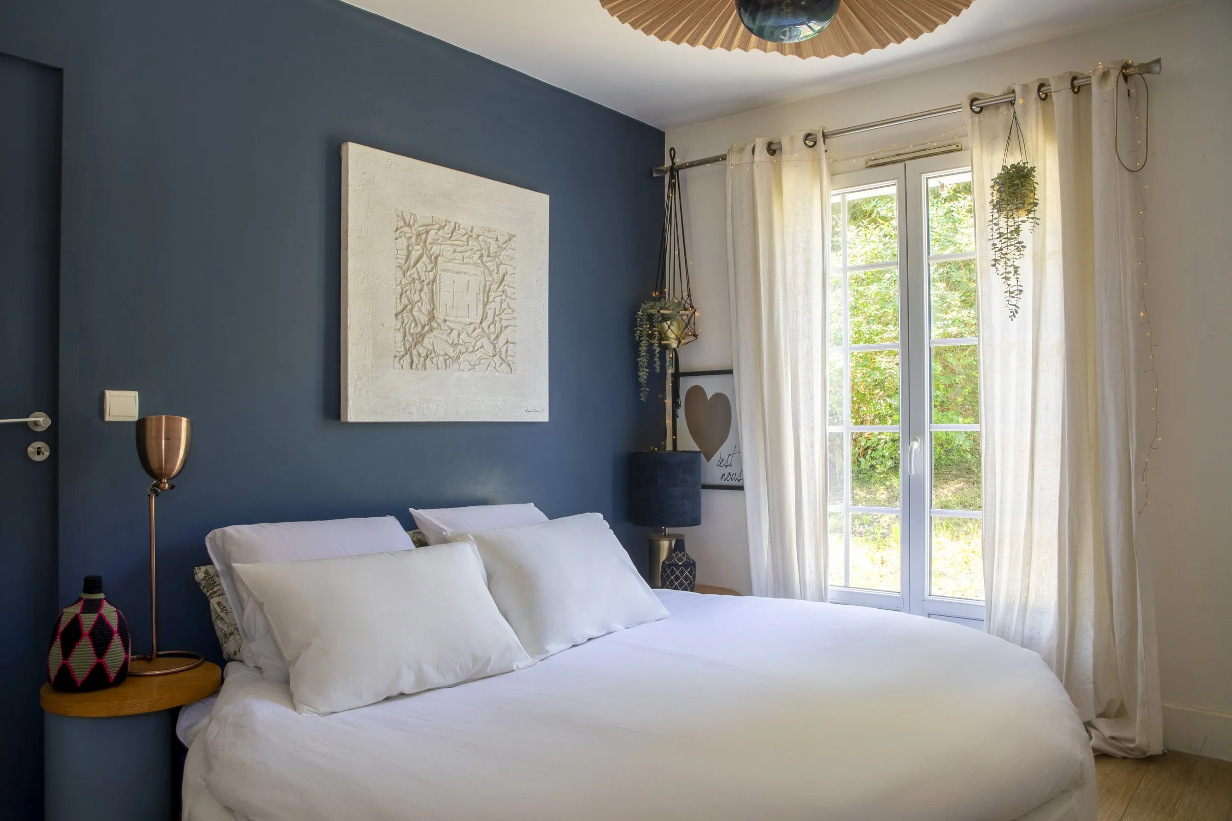 Chambre avec mur bleu derrière le lit, fenêtres avec rideaux blancs, décoration moderne, plantes suspendues, tableau blanc avec un cœur et une inscription, lampe en cuivre, lit avec draps blancs et coussins.