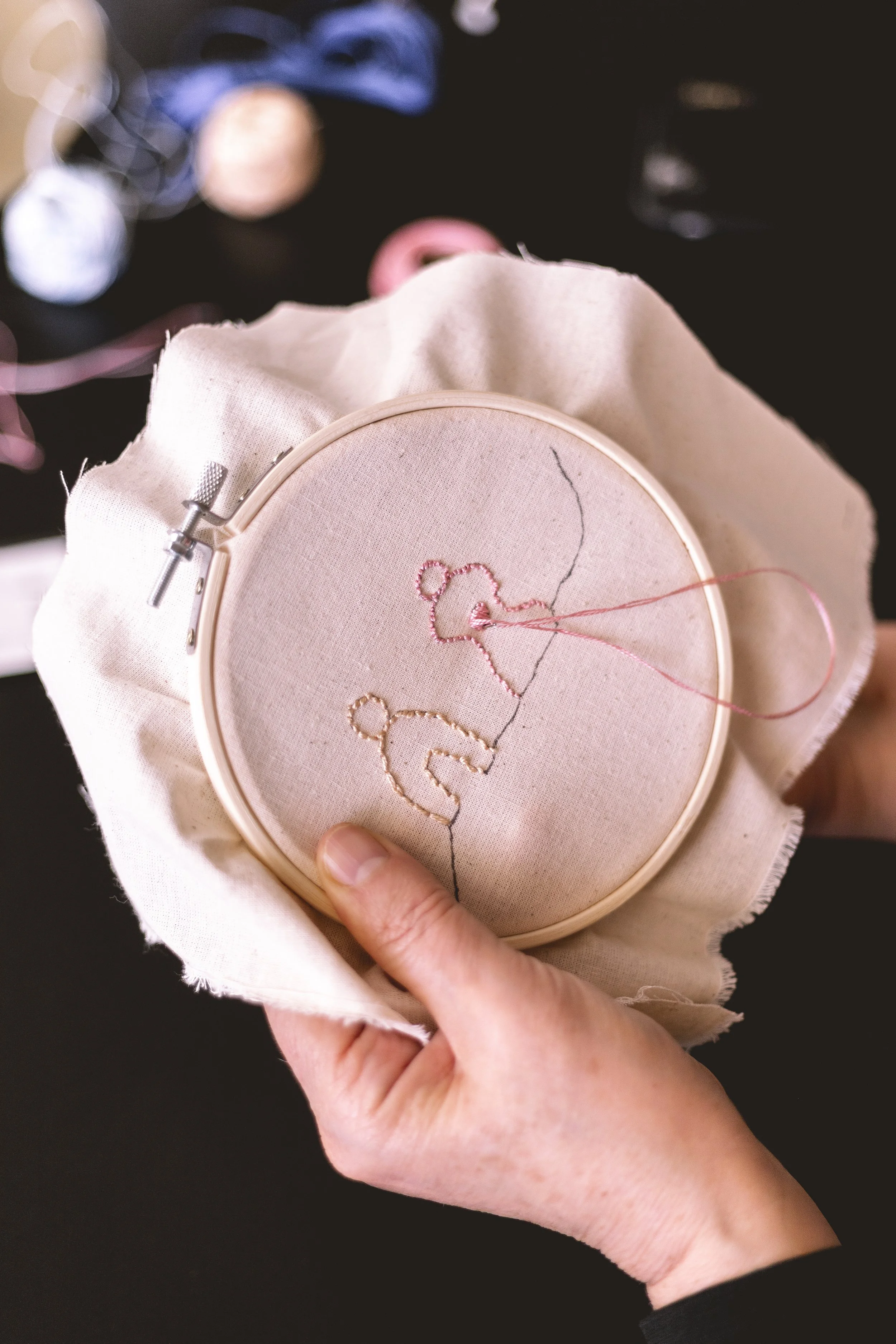 Une personne tient un cercle à broder avec la lettre « C » en fil rose, le cercle est fixé dans un cadre à broder, et la personne travaille sur une pièce de tissu blanc