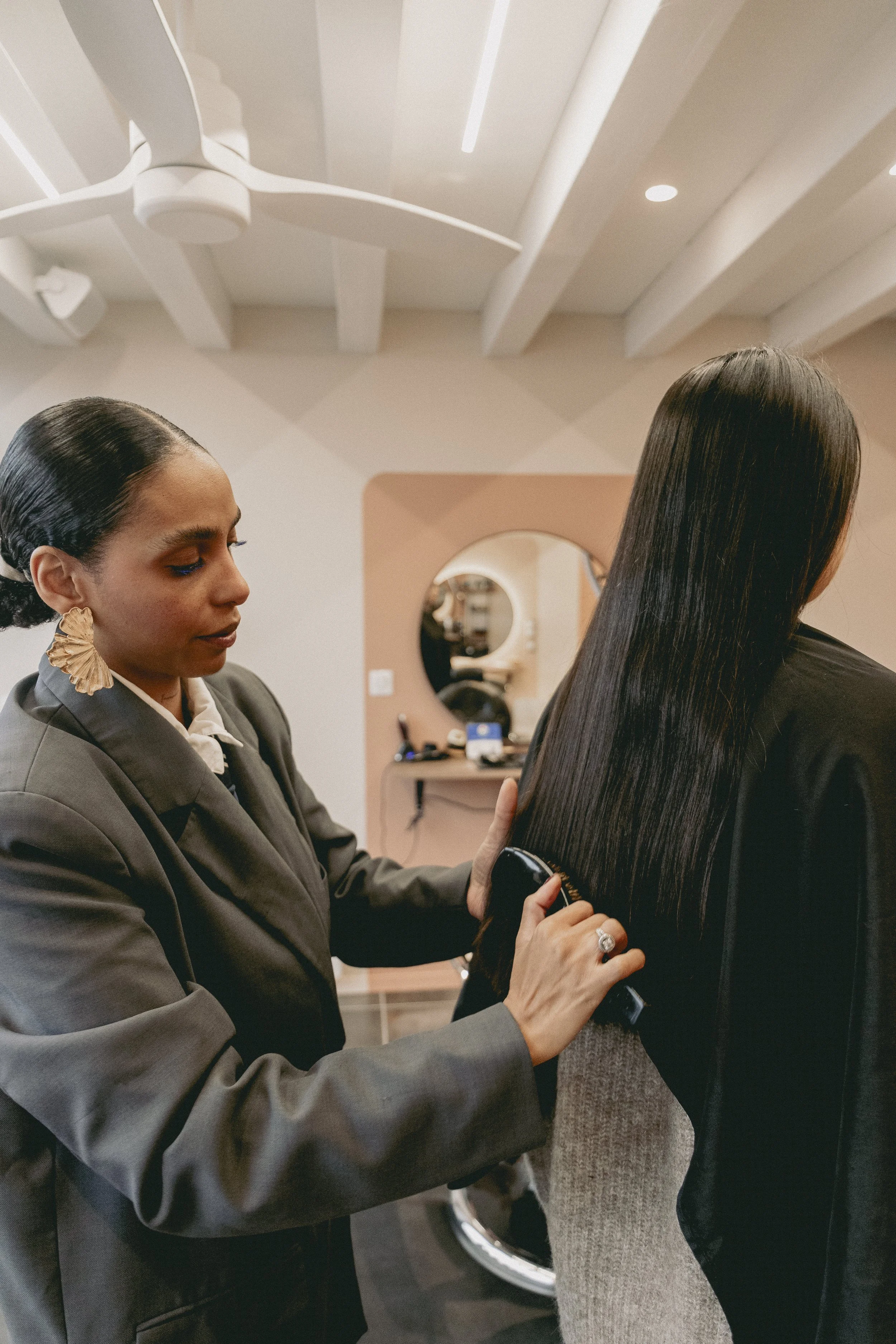 Une femme avec des cheveux noirs longs et brillants utilisant un sèche-cheveux sur une autre femme dont les cheveux sont noirs, raides et longs.