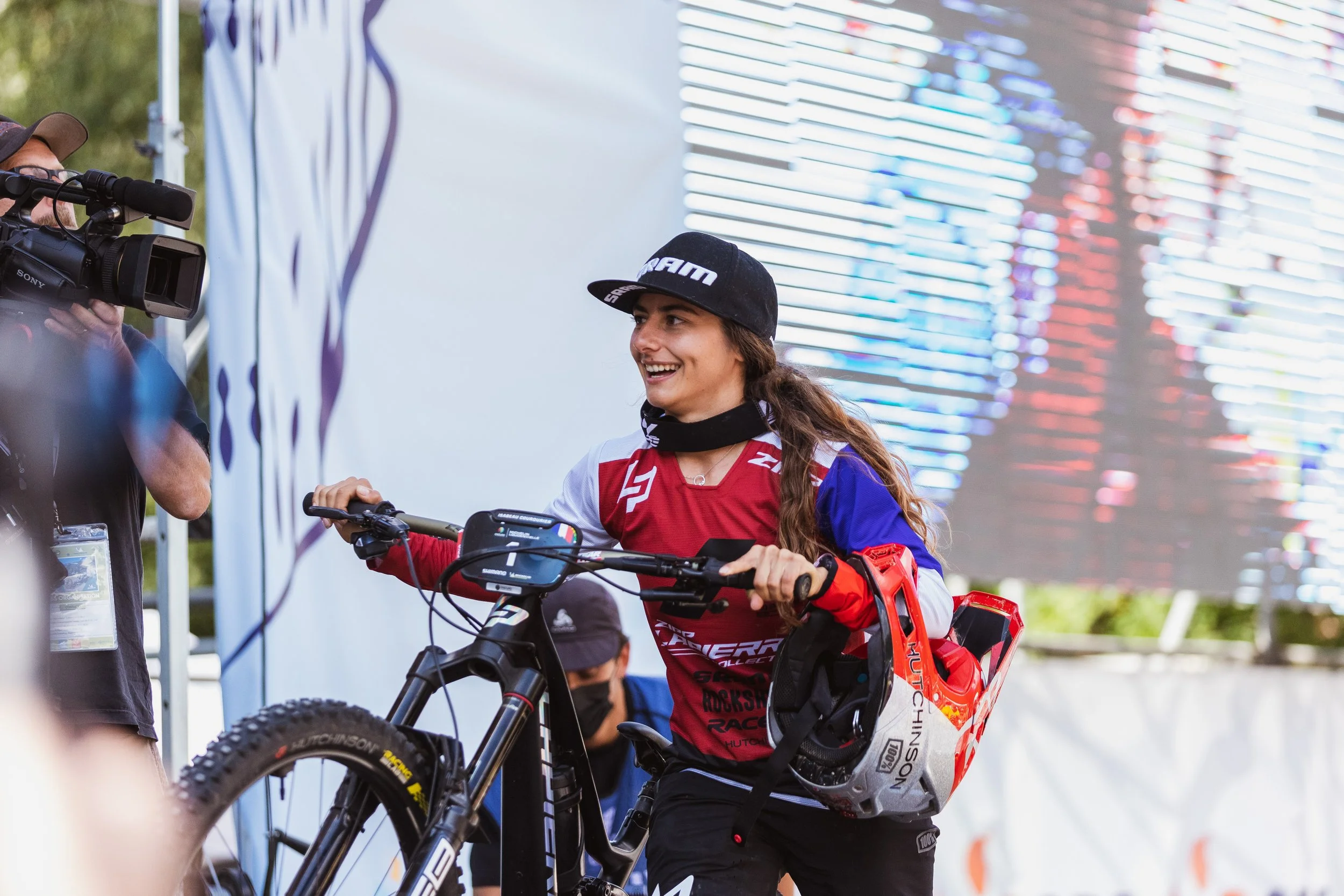 Une femme souriante en tenue de motocross, tenant un vélo tout-terrain lors d'une compétition ou d'un événement, avec un caméramans à sa gauche qui filme la scène et un décor coloré en arrière-plan.