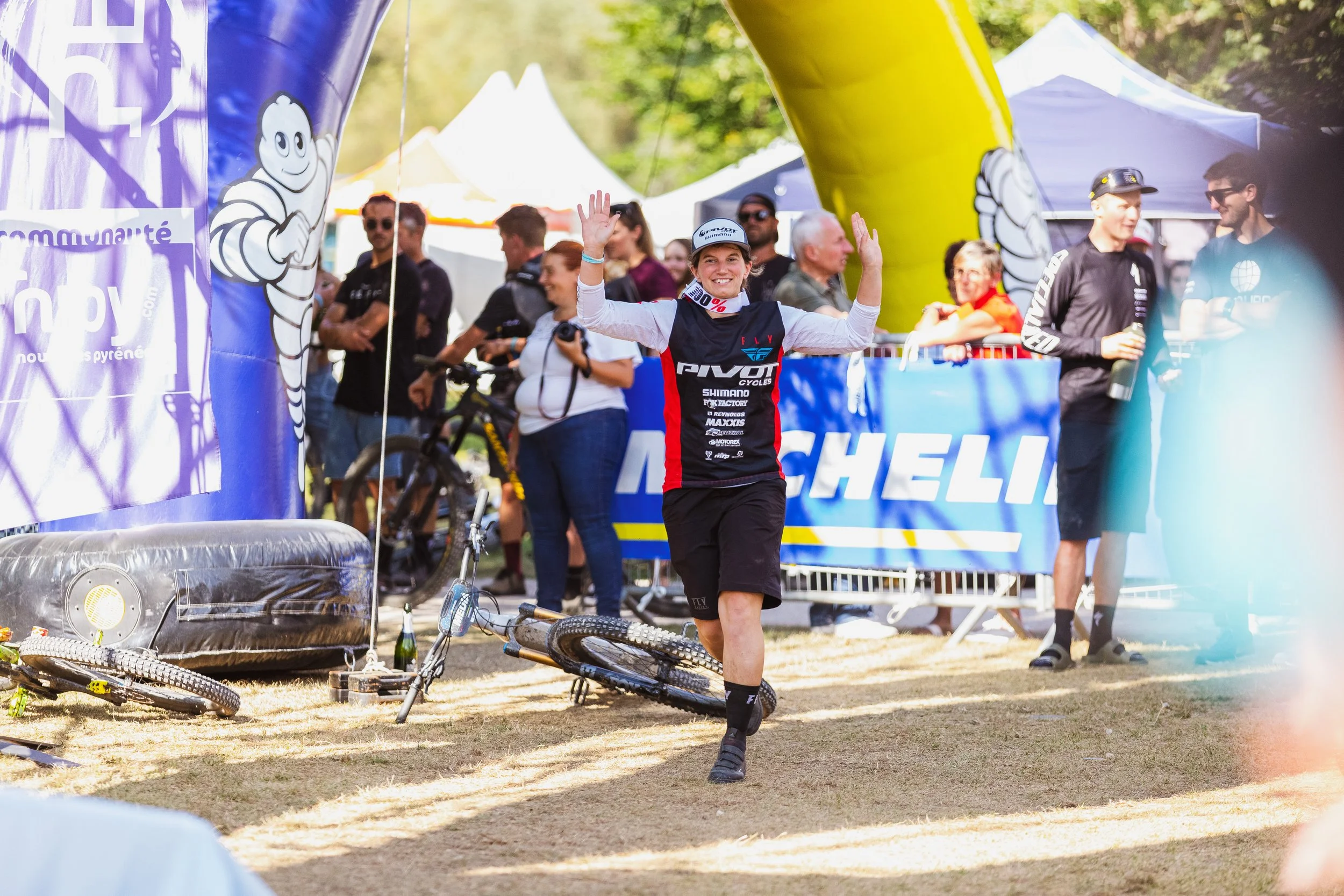 Une femme souriante en tenue de cyclisme franchit la ligne d'arrivée d'une compétition de VTT, entourée de spectateurs et d'organisations, sous un chapiteau bleu avec le logo Michelin.