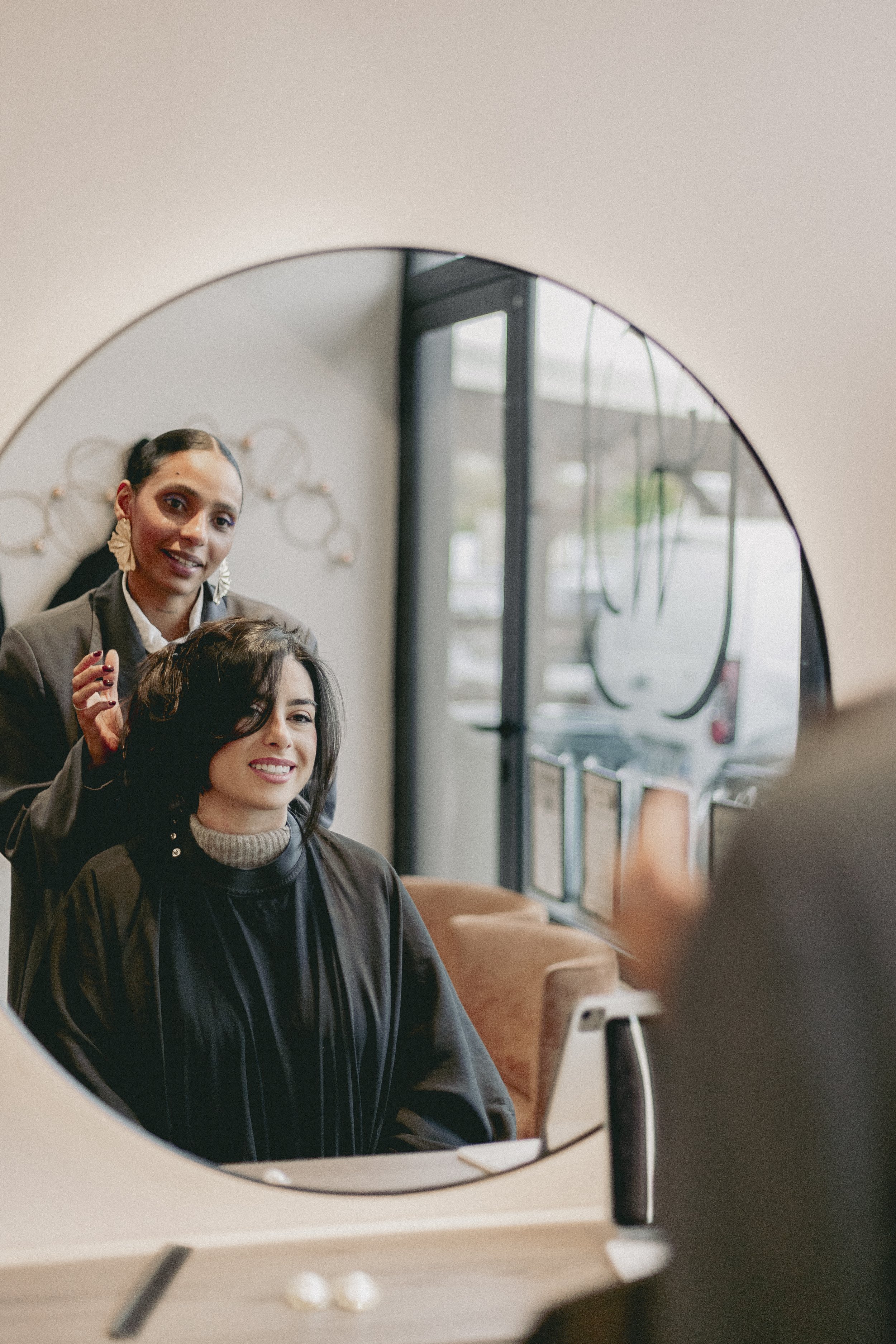 Une femme regarde son reflet dans un miroir rond, tandis qu'une autre femme se tient derrière elle et lui coiffe les cheveux, dans un salon de coiffure moderne avec de grandes fenêtres.