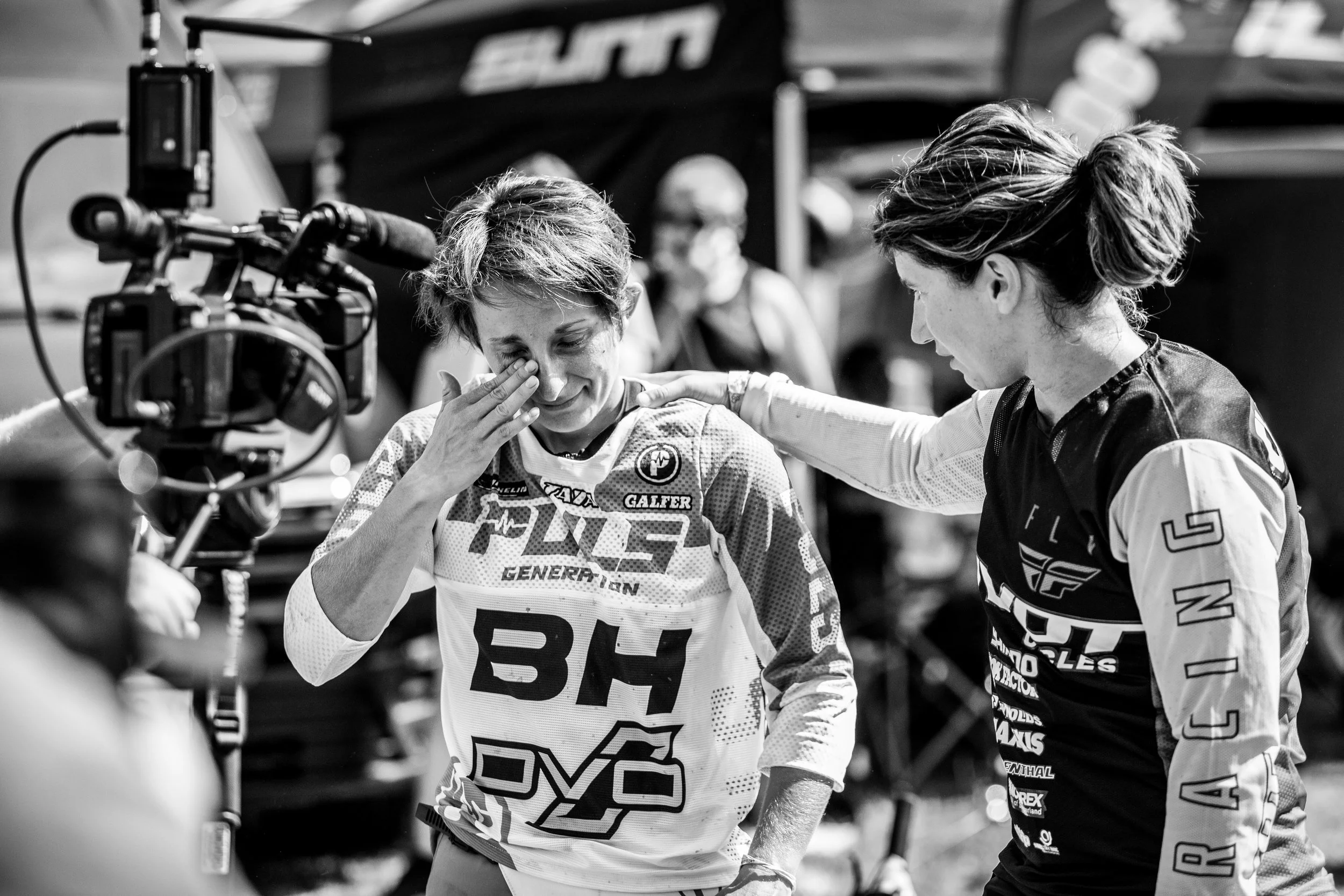 Deux femmes dans un environnement de course, probablement après une compétition. L'une d'elles, portant un maillot de course avec le numéro 35, semble émue ou en pleurs, tandis que l'autre lui touche l'épaule en signe de soutien. Il y a une caméra et