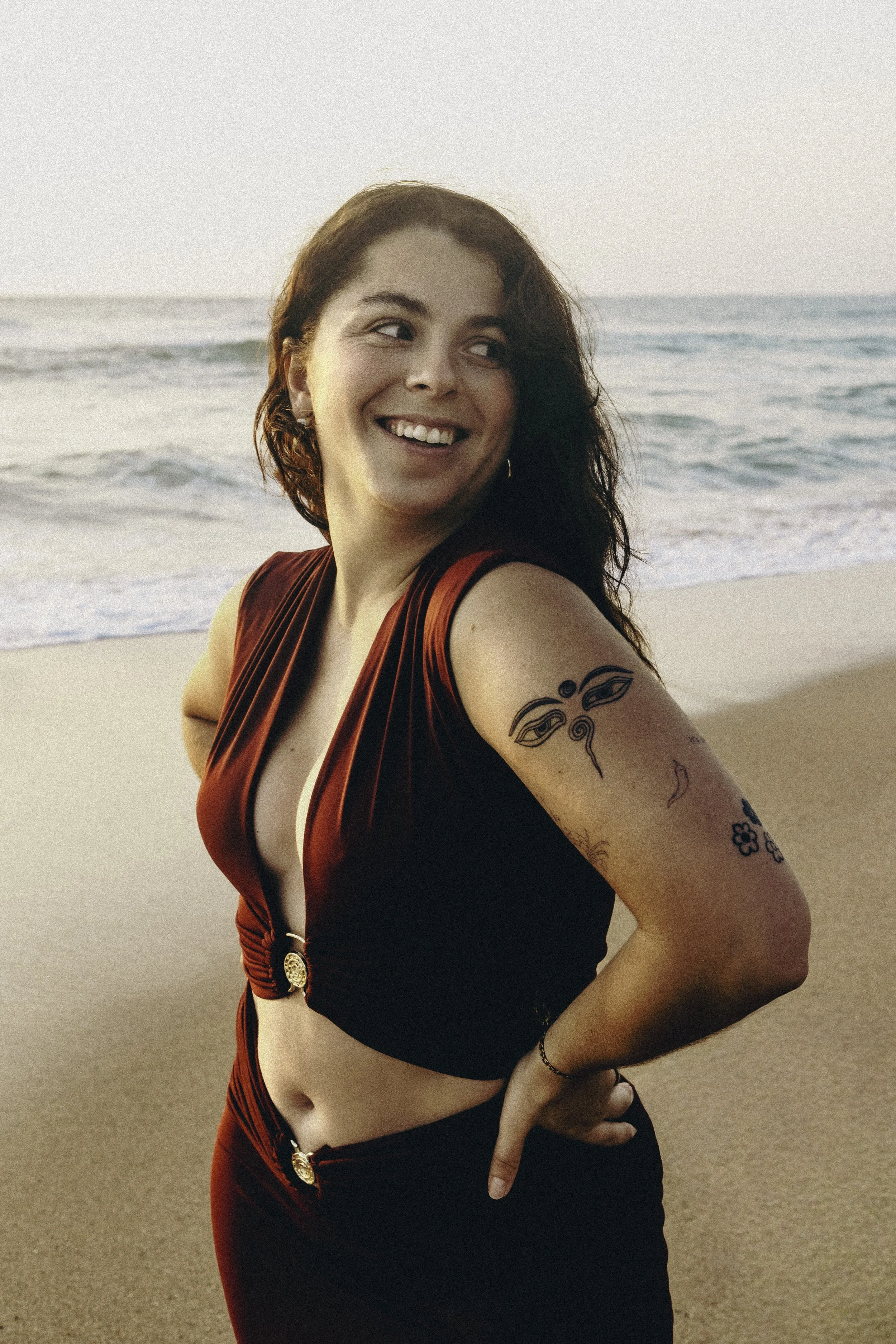 Une jeune femme souriante avec des tatouages sur le bras, portant une tenue rouge et noire, se tient sur une plage au coucher du soleil avec la mer en arrière-plan.