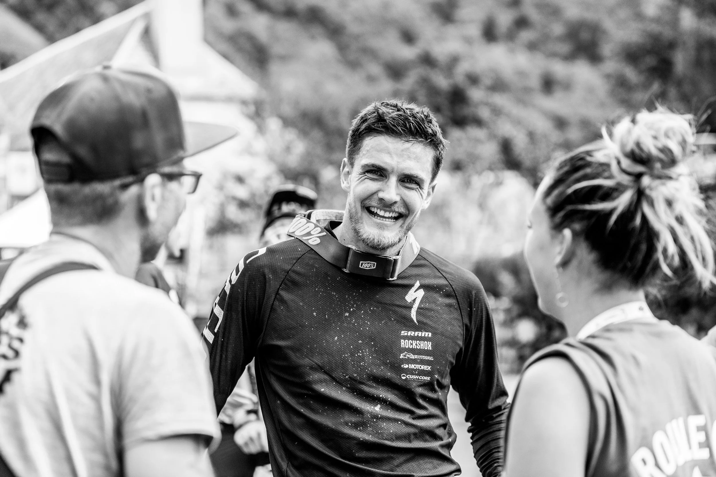 Groupe de personnes, dont un homme souriant en tenue de motocross, en conversation lors d'un événement en plein air.