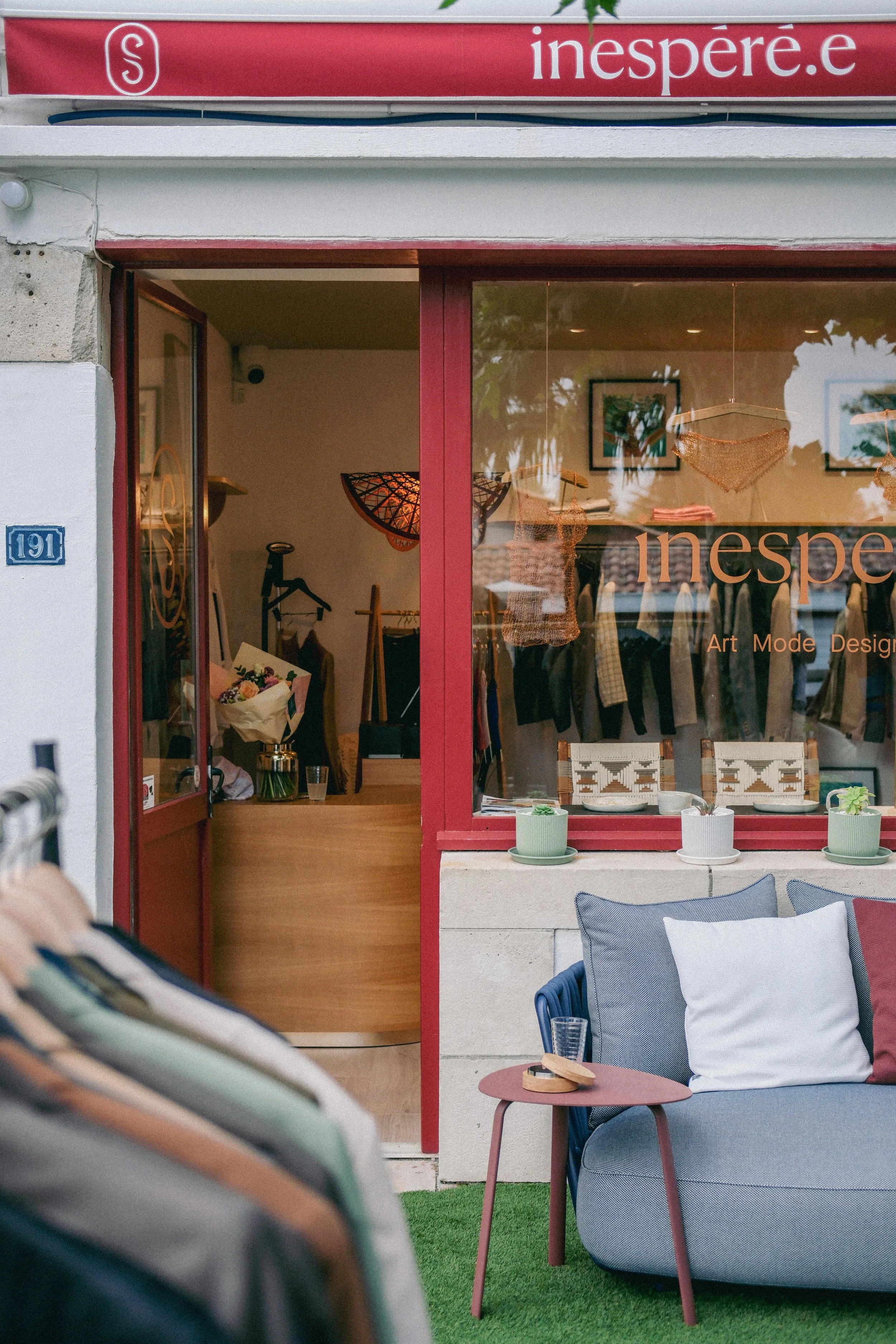 Vitrine d'une boutique avec objets de décoration et vêtements, reflet de l'extérieur dans la vitre, mobilier de jardin avec chaise en tissu bleu, table rose, coussins blancs et gris, plantes en pots, et panneaux peints à l'intérieur.