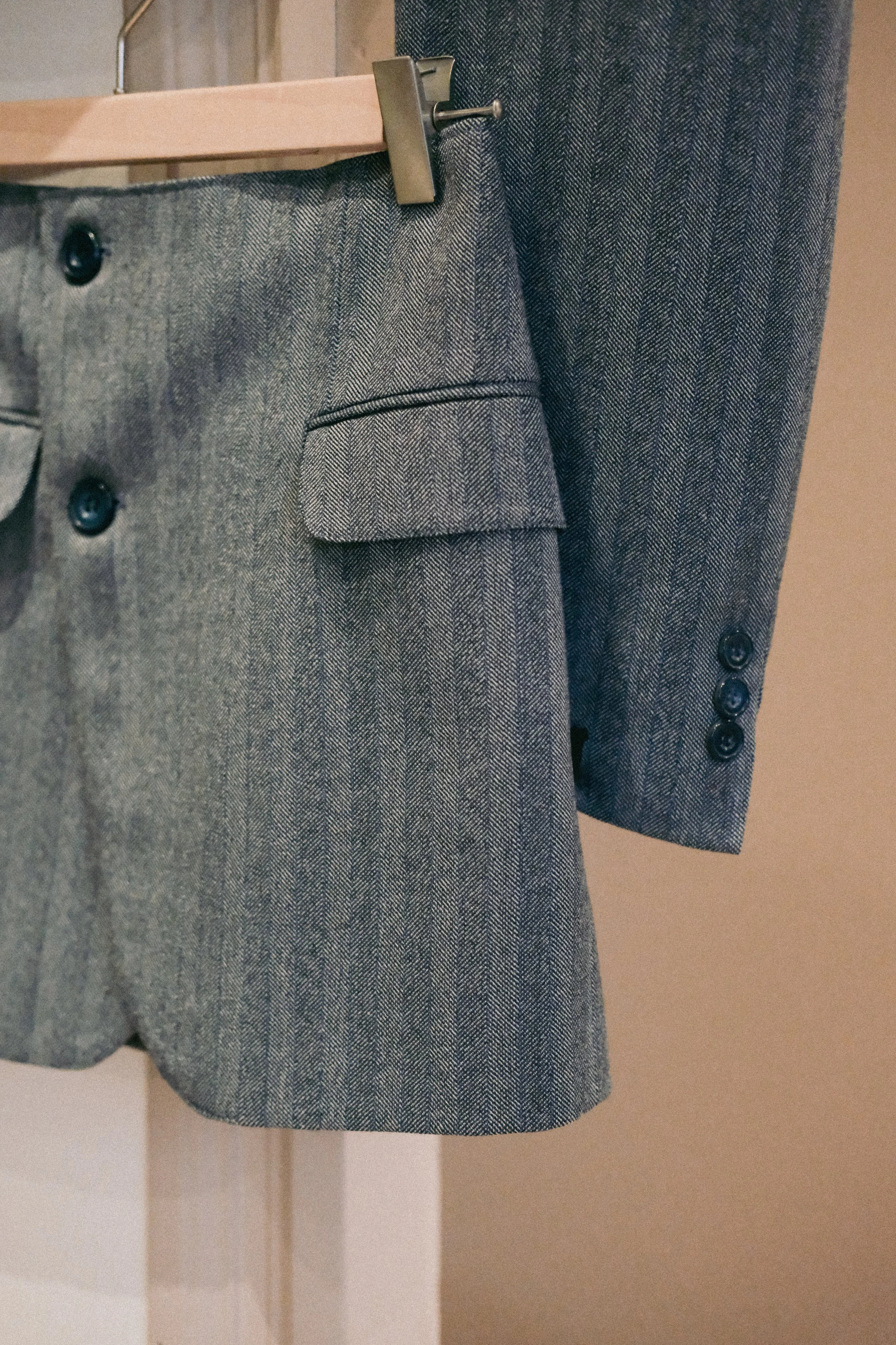 Costume gris à carreaux, comprenant une veste et une jupe ou pantalon, avec une poche et des boutons noirs, accroché à un portant.