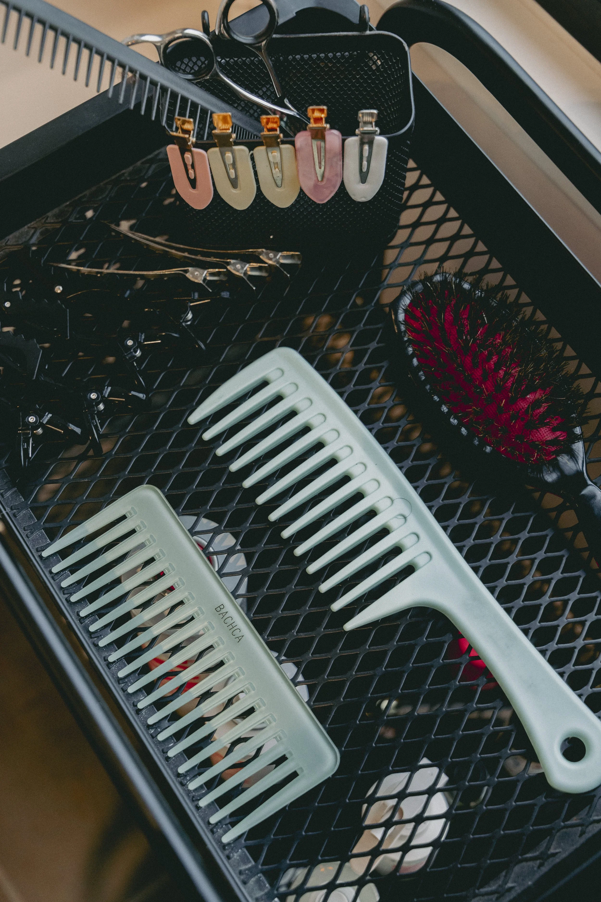 Panier noir contenant des accessoires de coiffure tels que des pinces à cheveux en différentes couleurs, un peigne blanc et une brosse à cheveux noire et rouge.