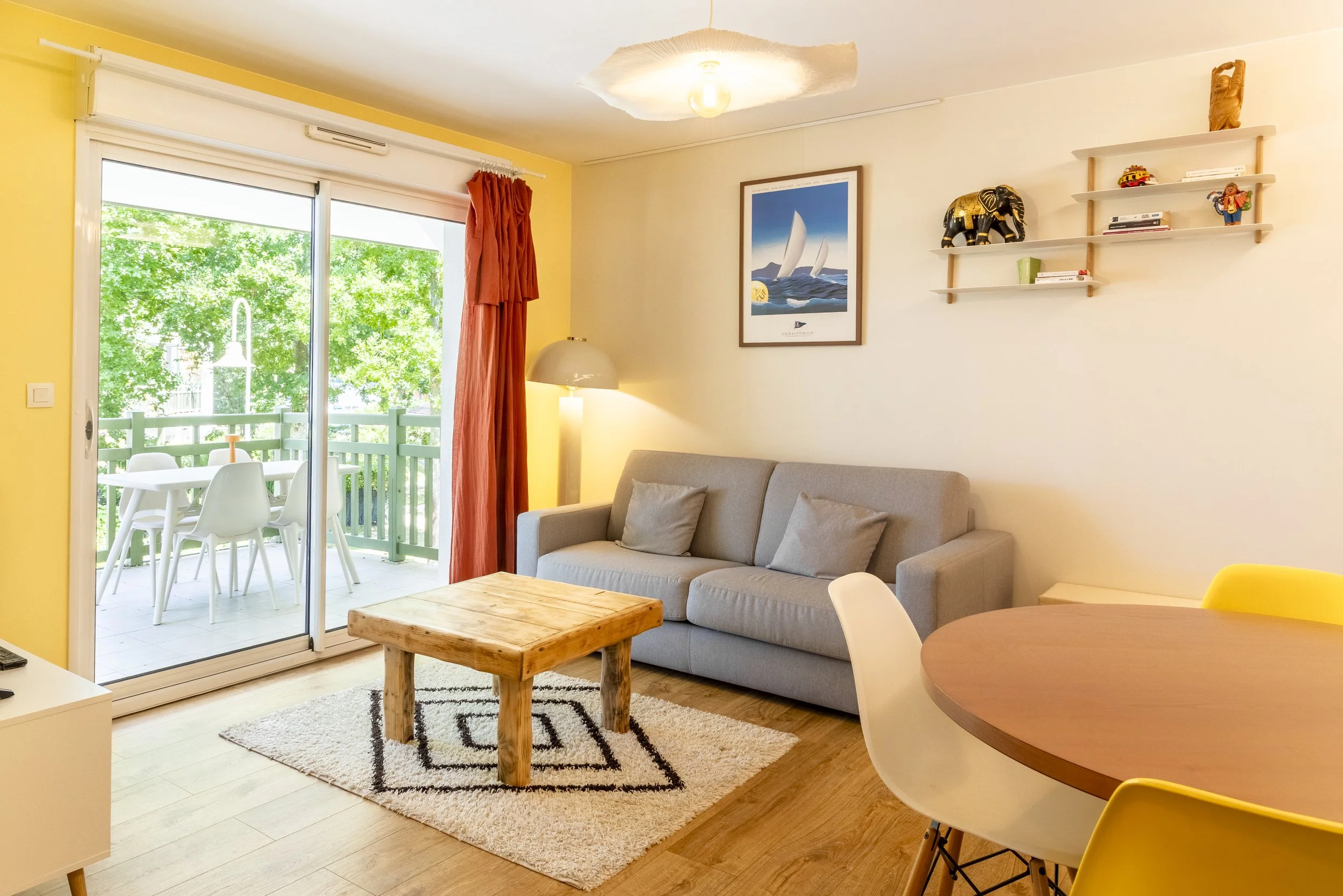 Salon avec canapé gris, tapis beige et table en bois brut, mur peint en blanc et jaune pâle, étagère en bois avec décorations et livres, tableau représentant des voiliers, porte-fenêtre donnant sur balcon avec table et chaises en plastique blanc, rideaux rouges, lampe sur pied, et partie de la cuisine avec table ronde et chaises jaunes