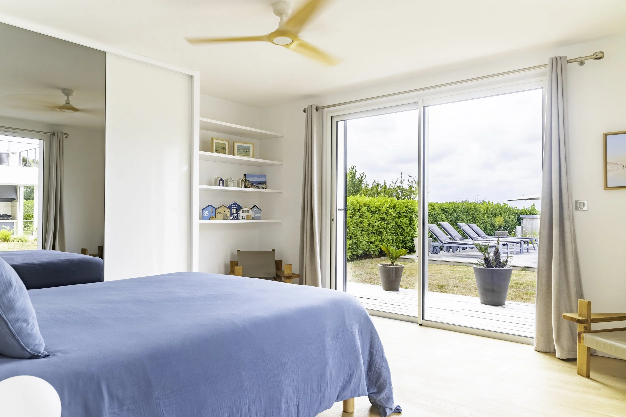 Une chambre avec un lit en drap bleu, une grande fenêtre donnant sur un jardin avec des chaises longues, et des étagères décoratives